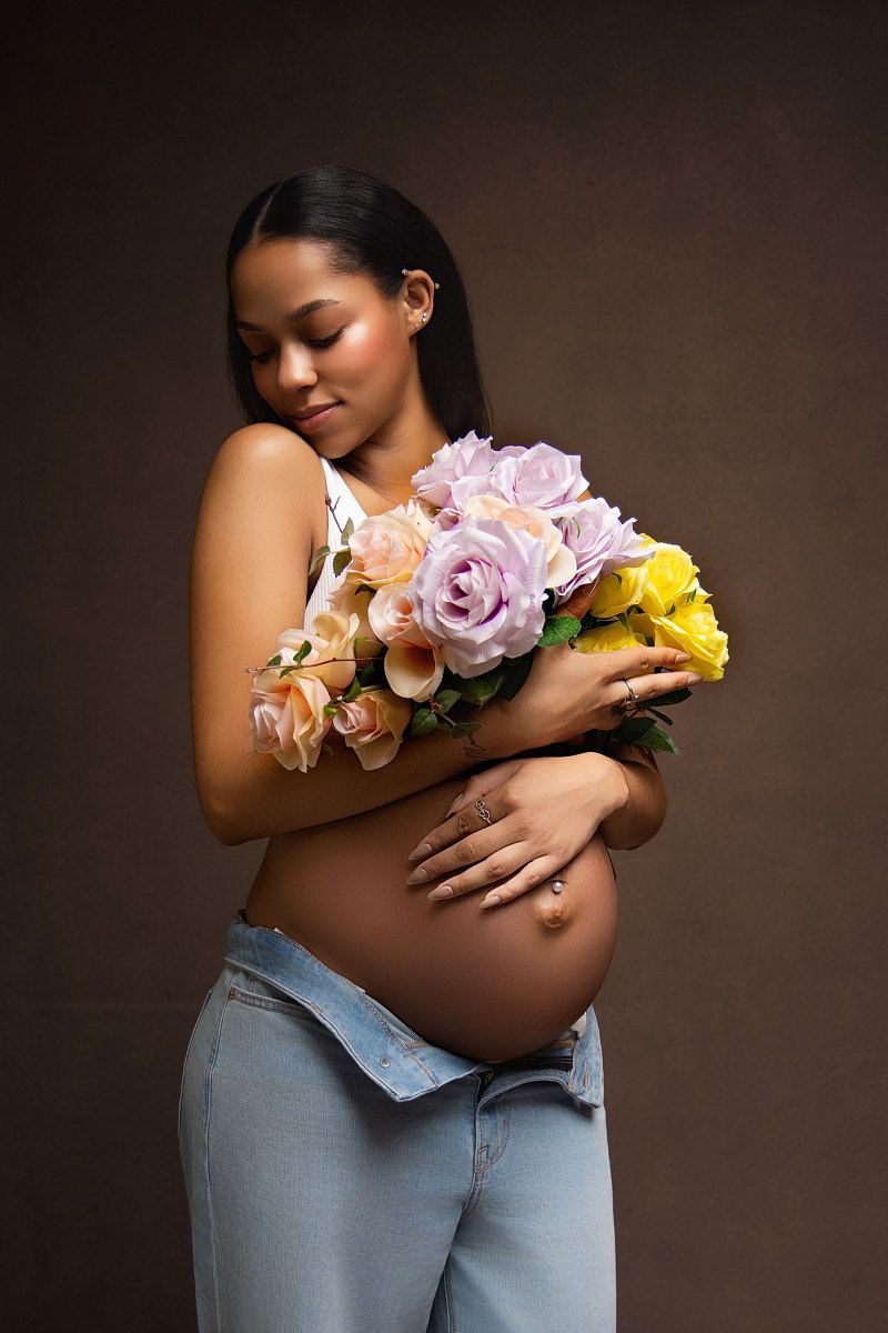 Beautiful maternity portrait with person holding pink and yellow roses against dark background wearing jeans.