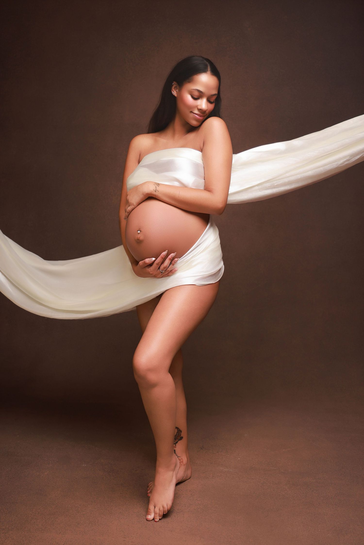 A pregnancy photoshoot in dark brown tones shows an intimate silhouette draped in flowing white fabric.