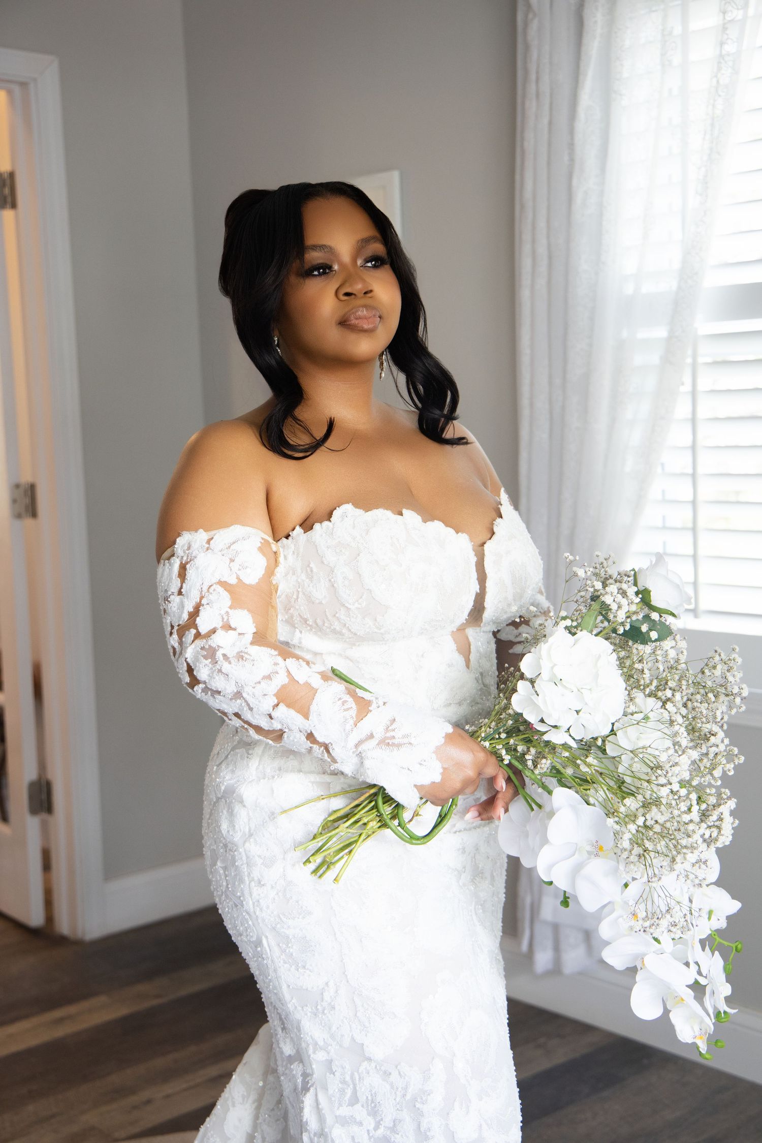 A bride in an off-shoulder lace wedding gown holds an elegant bouquet of white flowers and baby's breath.
