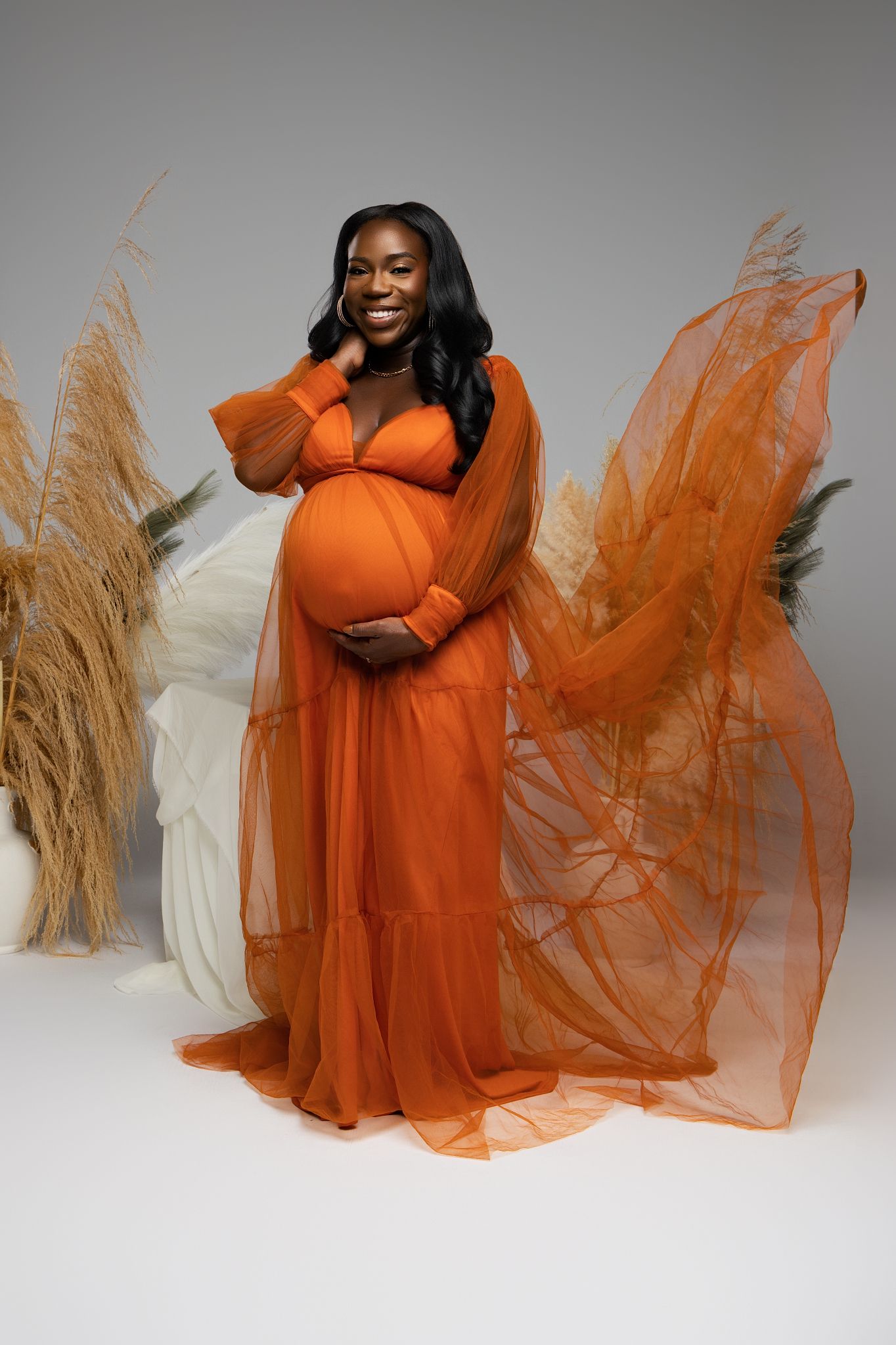 Solo maternity portraits in flowing orange gown with pampas grass backdrop in studio setting.