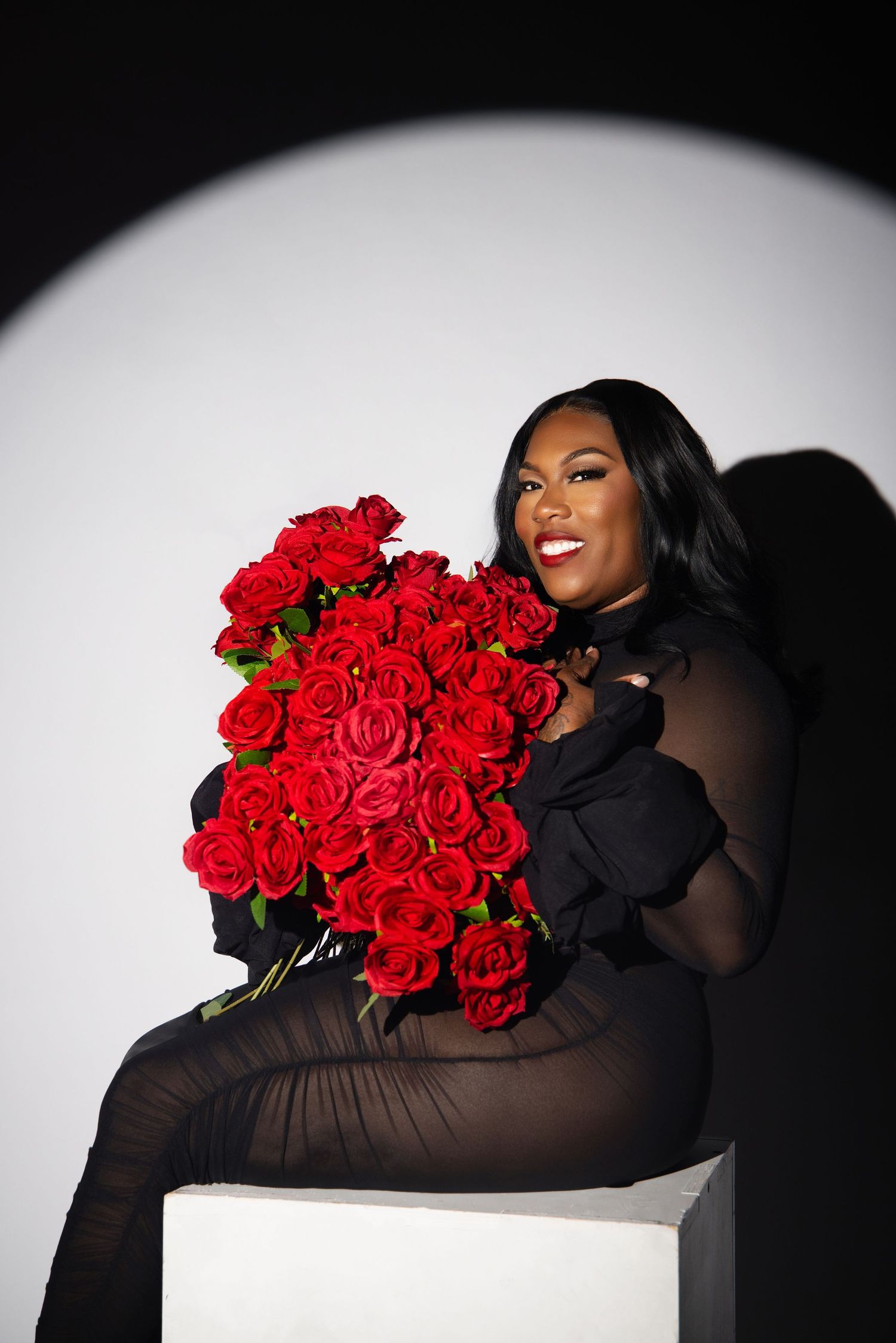 Model wearing black sheer dress poses on white cube with vibrant bouquet of red roses against dark background.