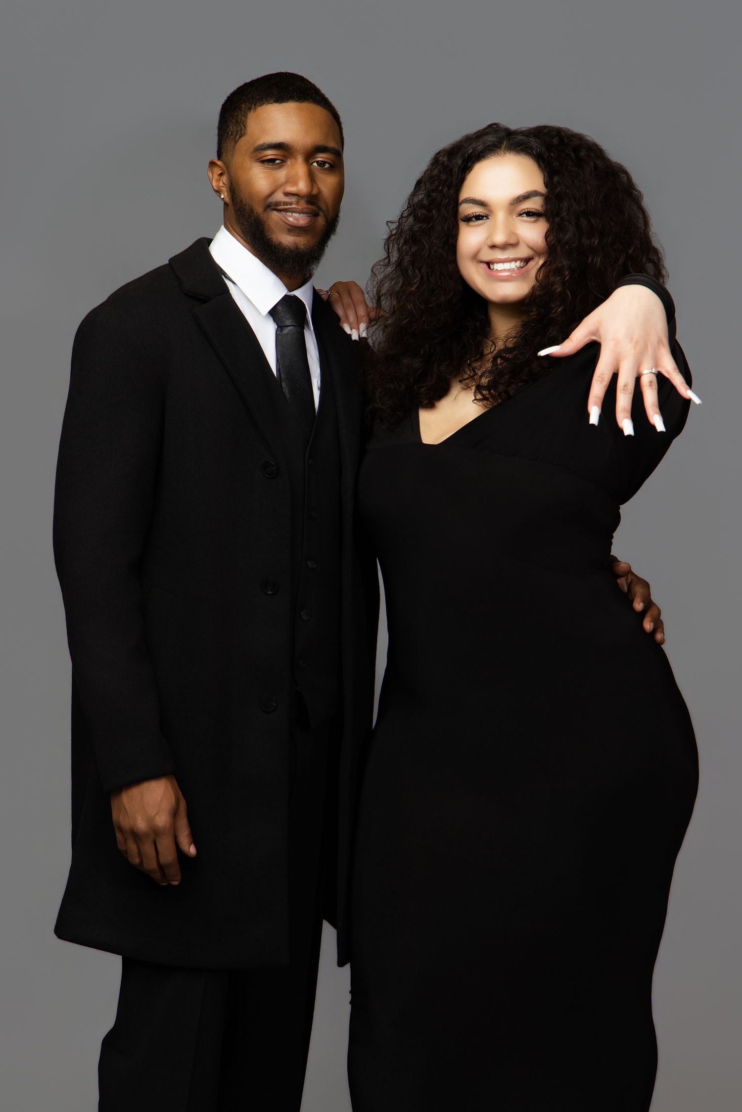 A couple in formal black attire poses together against a gray background for an elegant portrait.