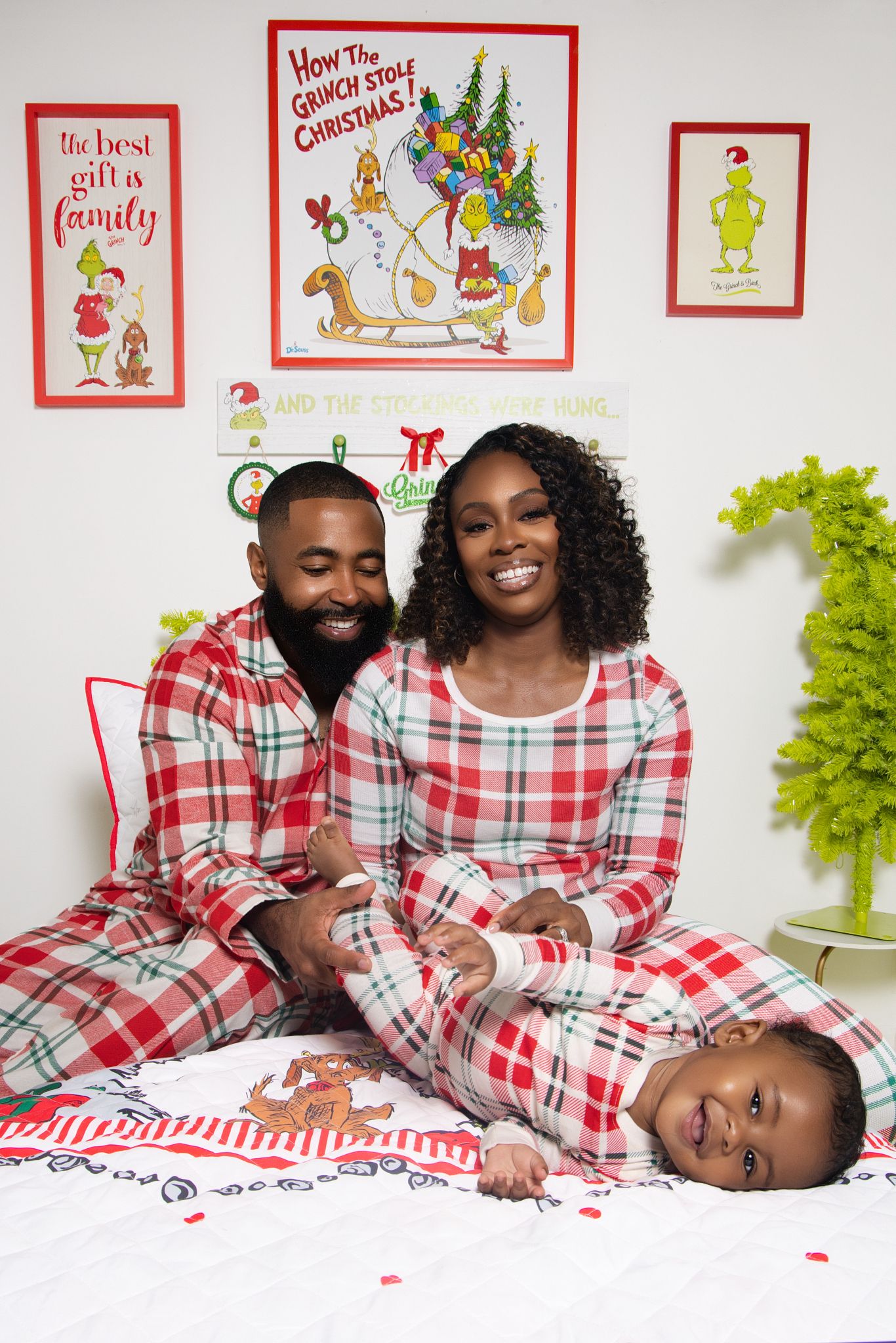A family wearing matching red and white plaid Christmas pajamas poses together on a white background with holiday decor.