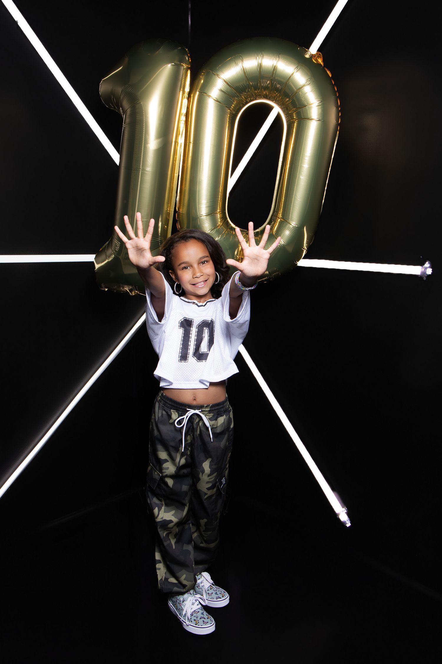 A young girl celebrates her 10th birthday holding gold number balloons, wearing a jersey and camo pants.