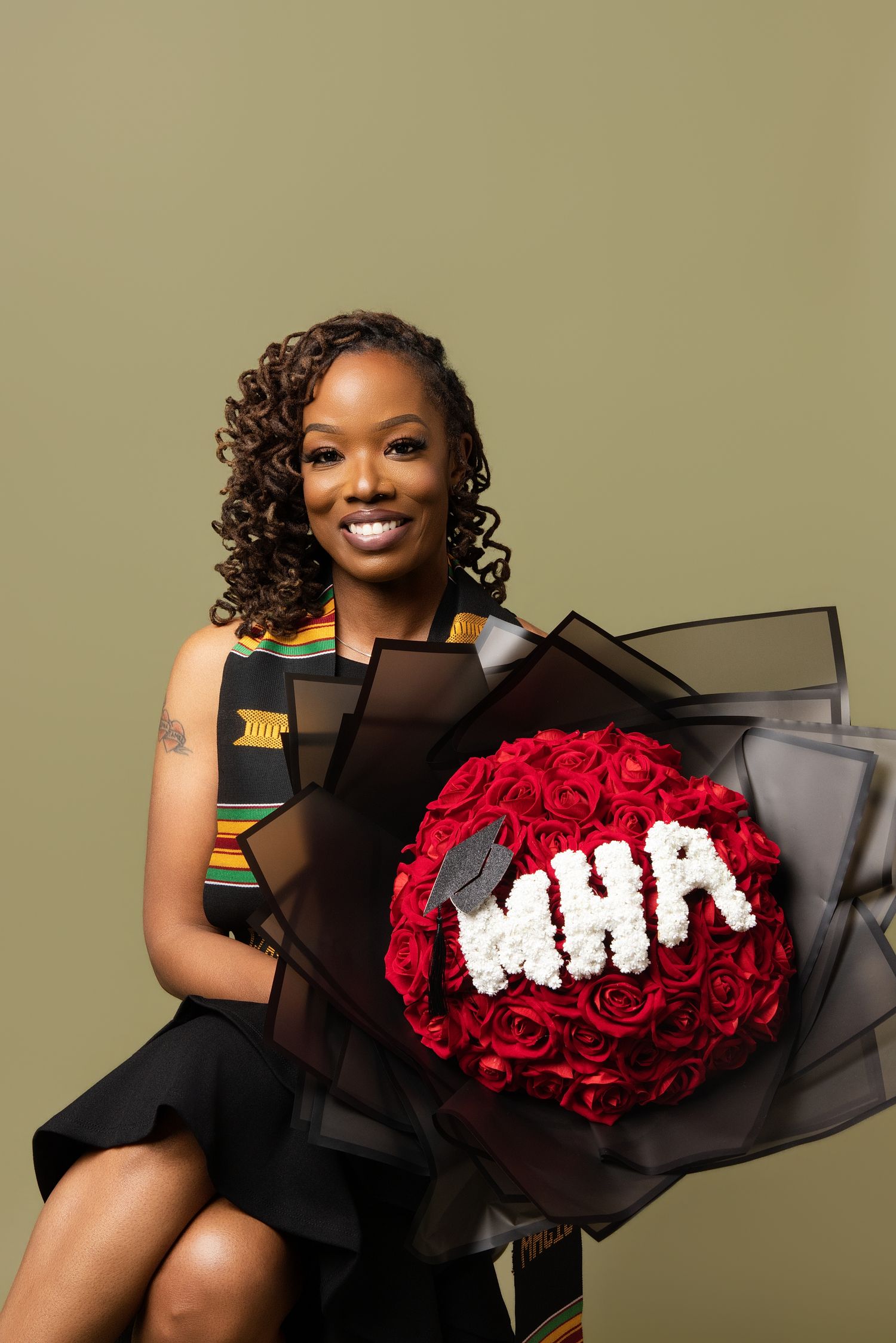 Smiling graduate holding a bouquet of red roses spelling MHA, wearing a kente cloth stole, celebrating her master's degree.
