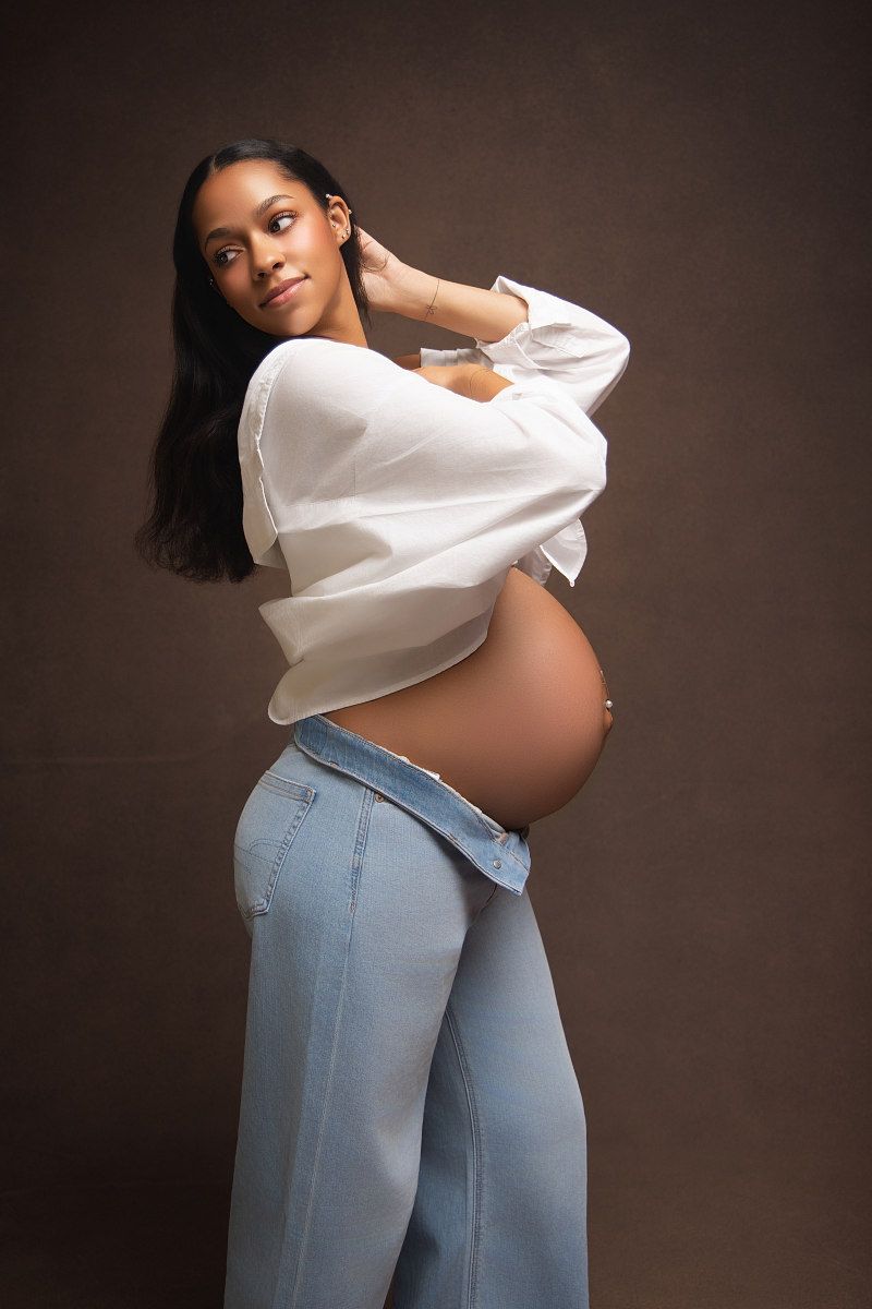 A pregnant person in light blue jeans and white top poses against a dark brown background in a maternity photoshoot.