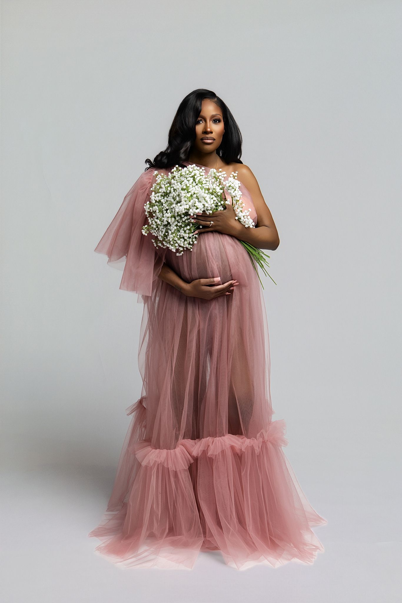 A woman in a flowing dusty pink tulle gown holds a bouquet of baby's breath flowers in a studio setting.