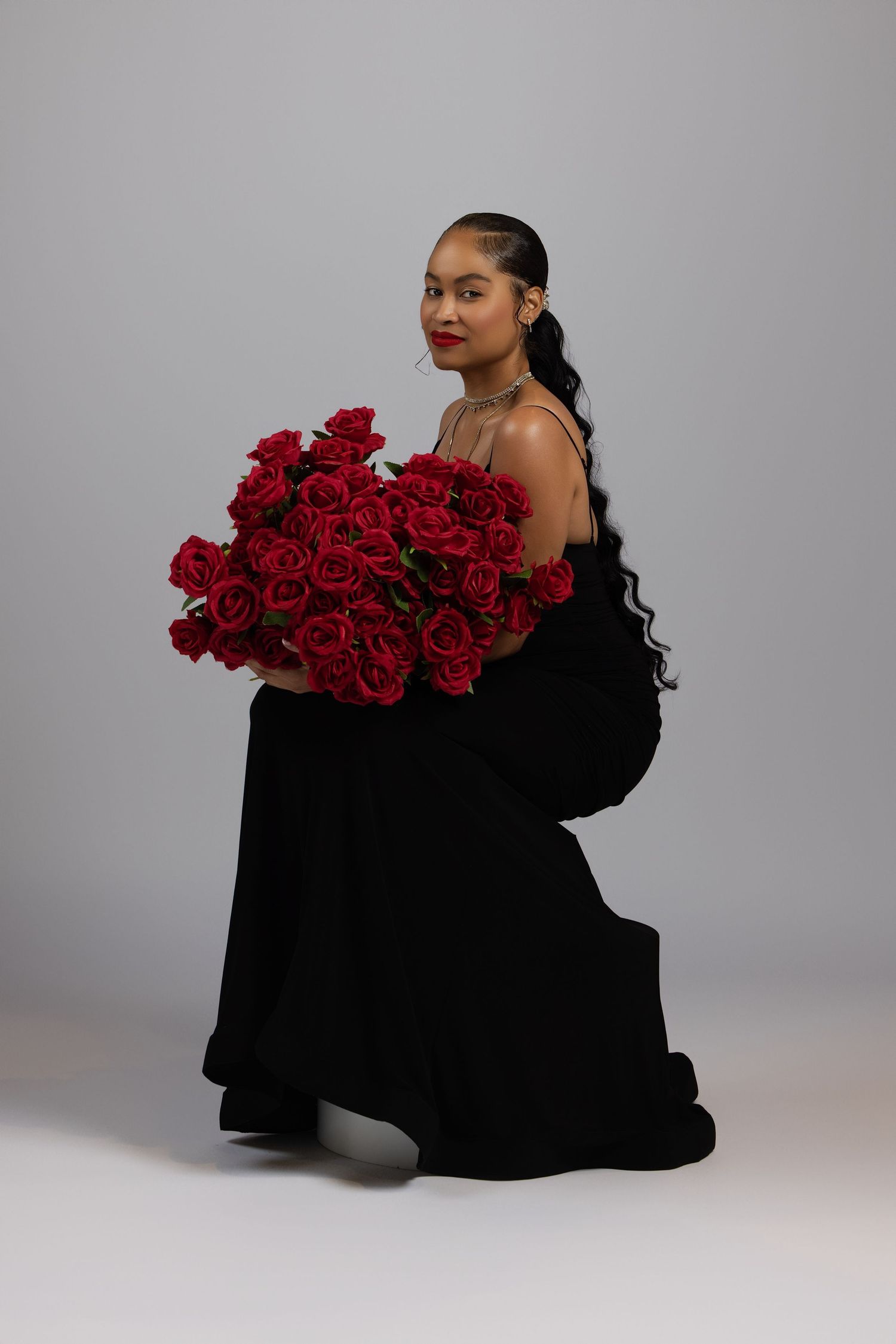 Figure in flowing black dress poses elegantly with vibrant red roses against light gray backdrop in fashion photoshoot series.