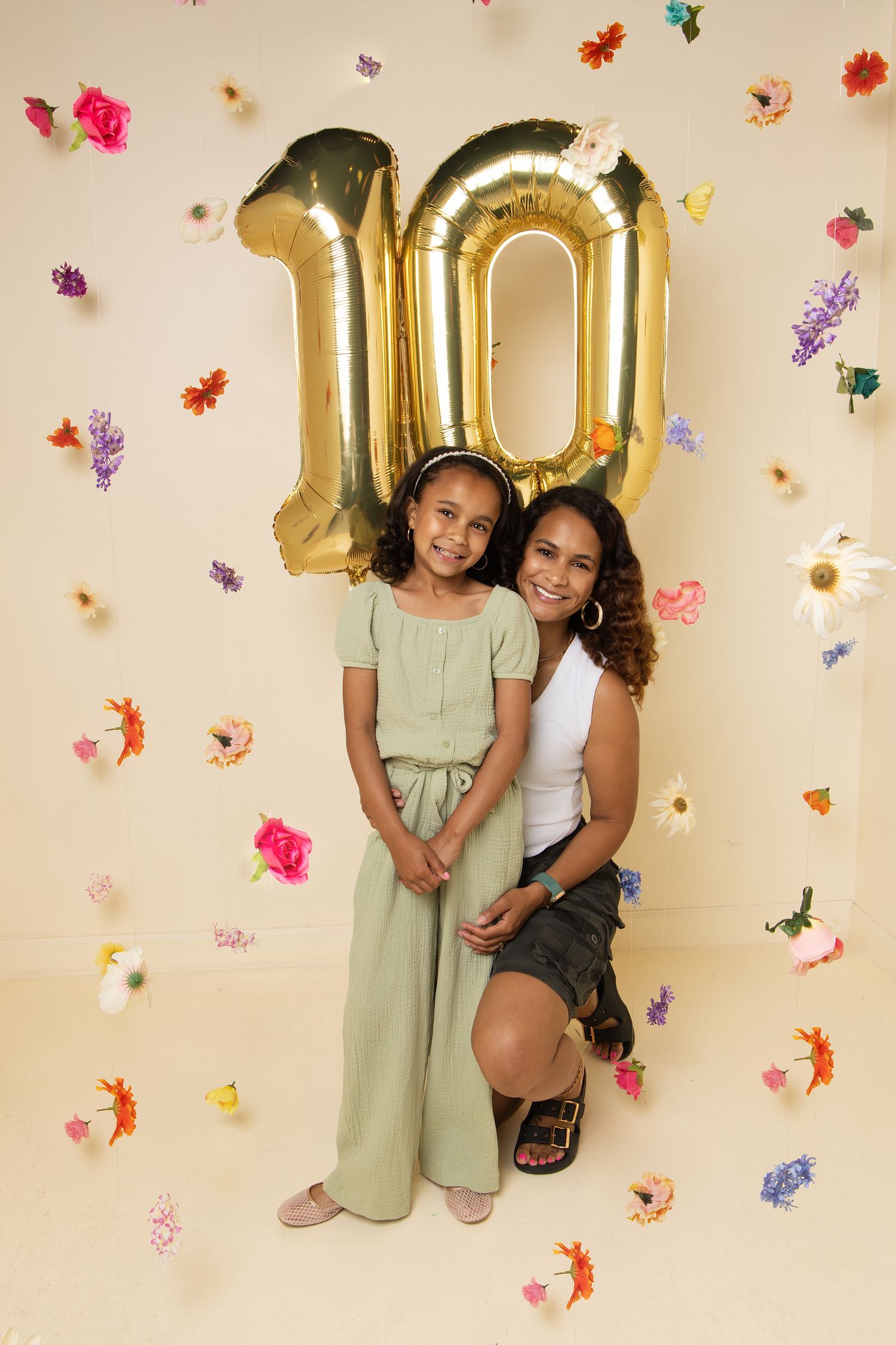 A woman and young girl pose together under gold number 10 balloons surrounded by colorful flowers on a cream background.
