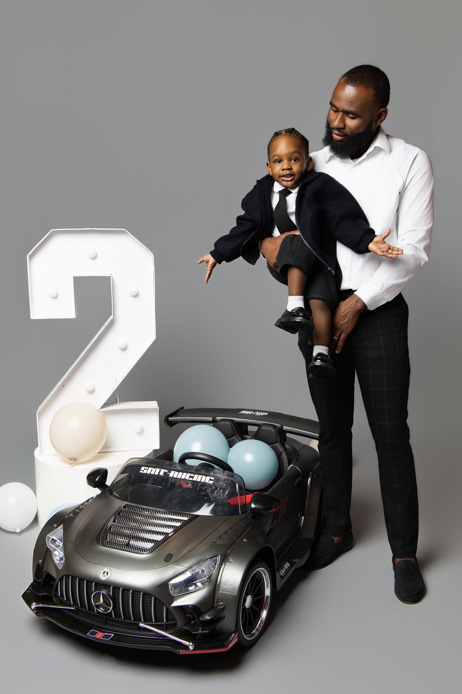 A toddler and adult pose with a large number 2 prop and a black toy Mercedes AMG GT ride-on car against a gray background.