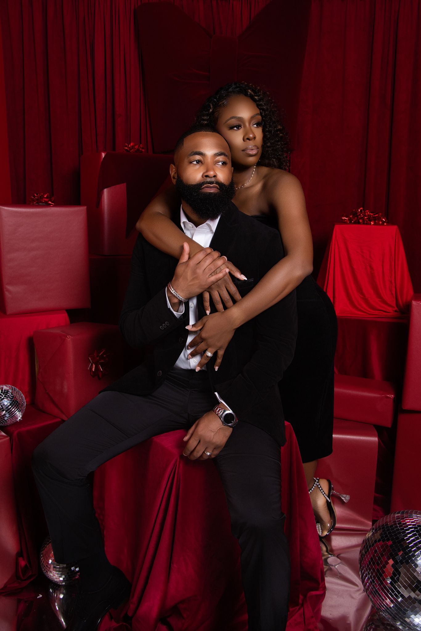 Elegant couple in formal black attire pose intimately against red velvet backdrop with disco balls and dramatic lighting.