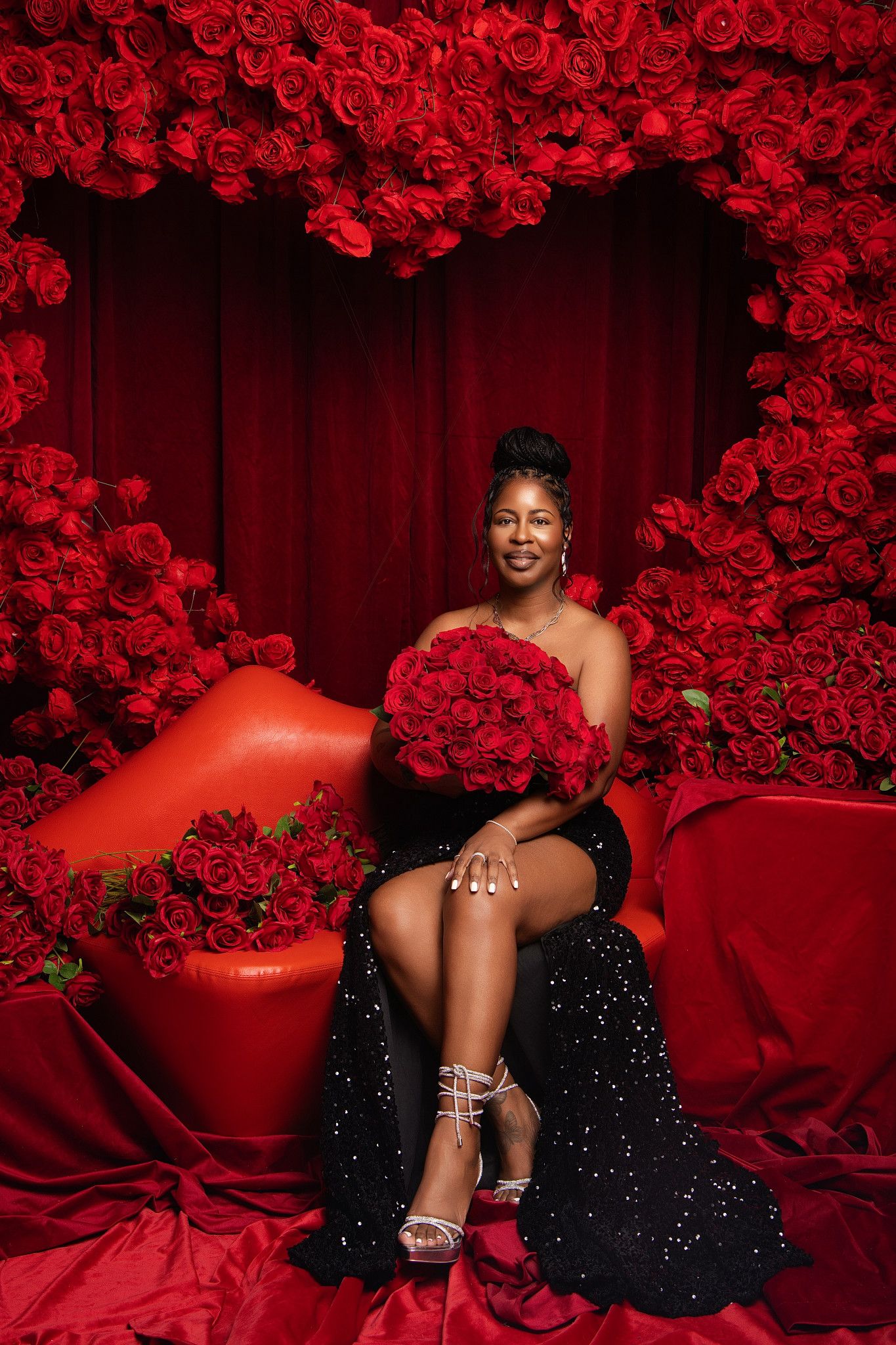 Artistic boudoir photoshoot with dramatic red roses and black sequin dress against luxurious red backdrop.