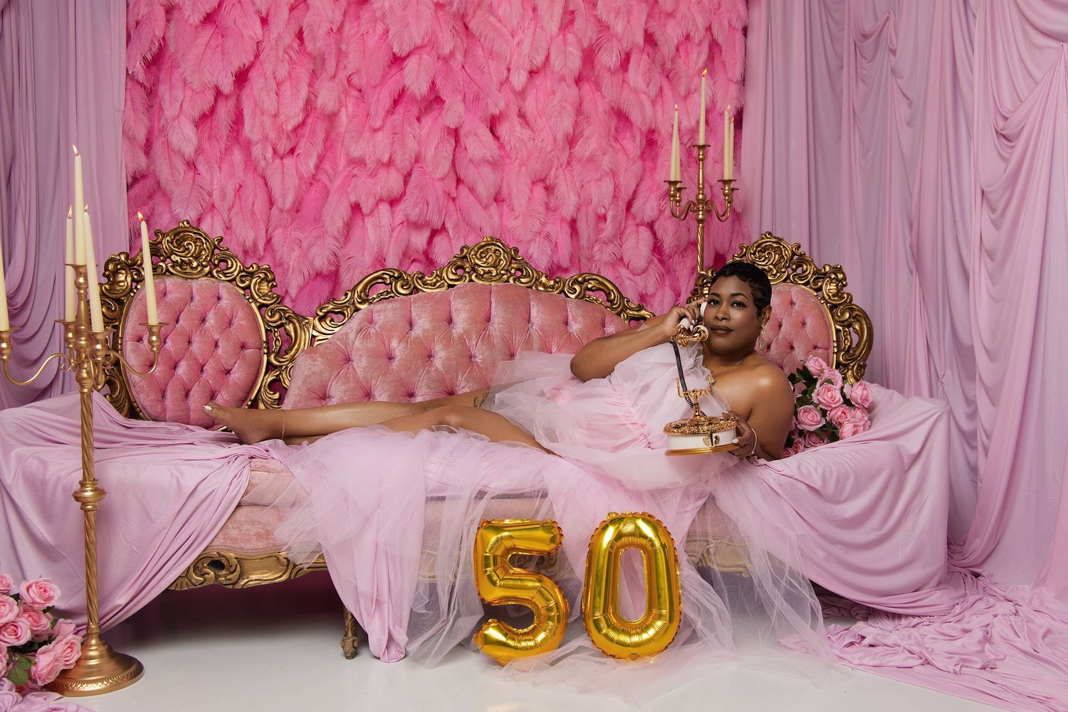 Woman in pink gown celebrating 50th birthday on ornate gold bed with pink floral backdrop and gold balloon numbers.