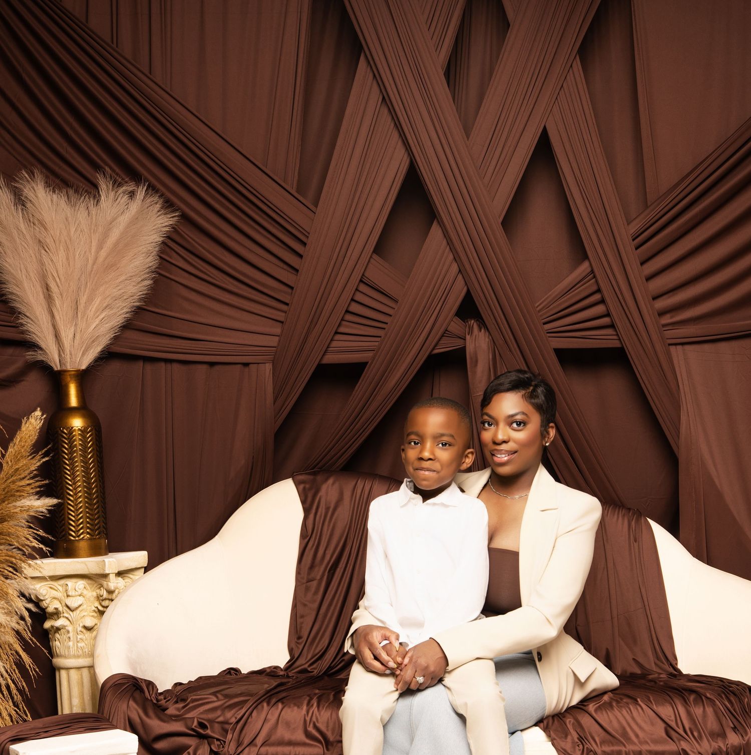 Two people in elegant white attire embrace in front of a textured brown fabric backdrop and decorative elements.