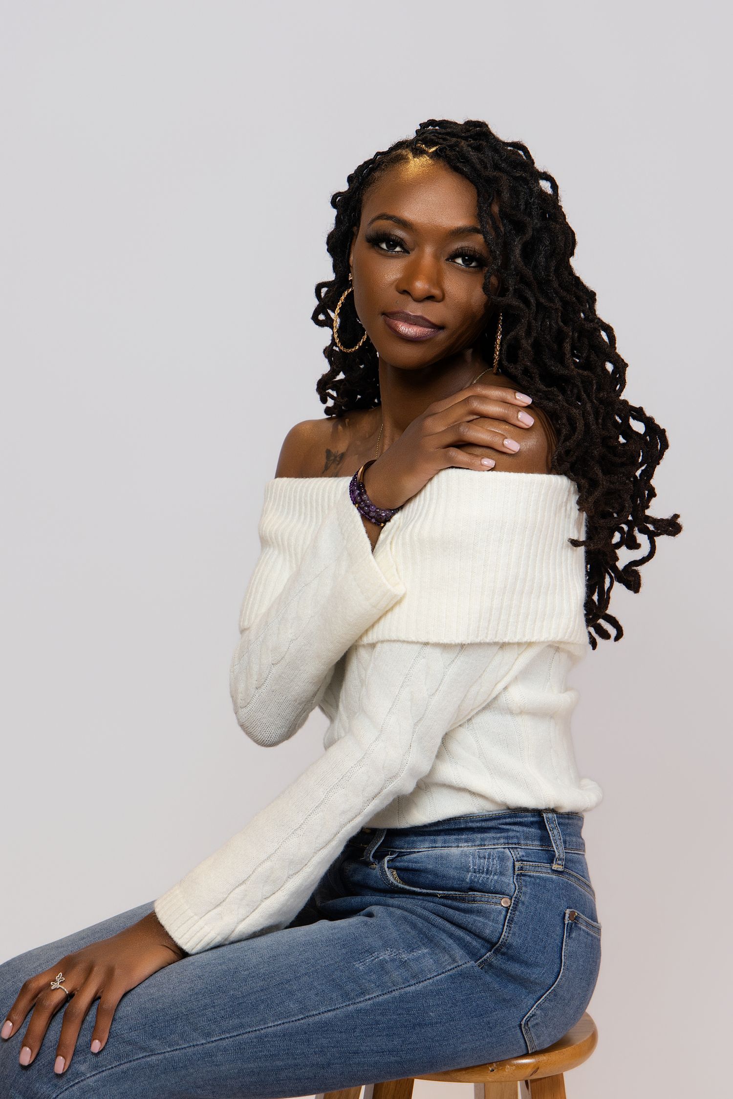 A person in a white knit sweater and blue jeans poses against a light gray background in a fashion photoshoot series.