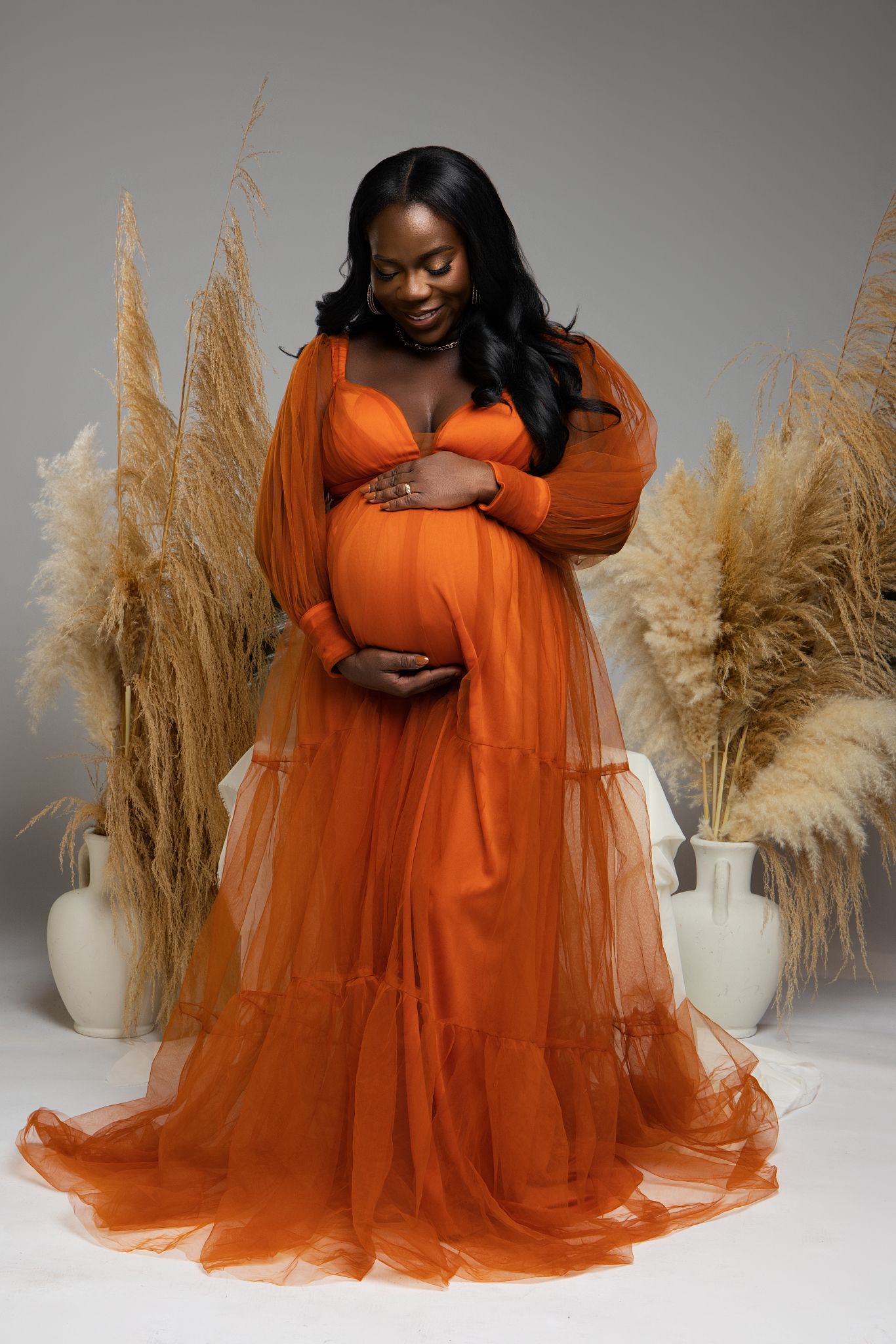 A maternity photo featuring a flowing burnt orange tulle gown against a backdrop of pampas grass and white vases.
