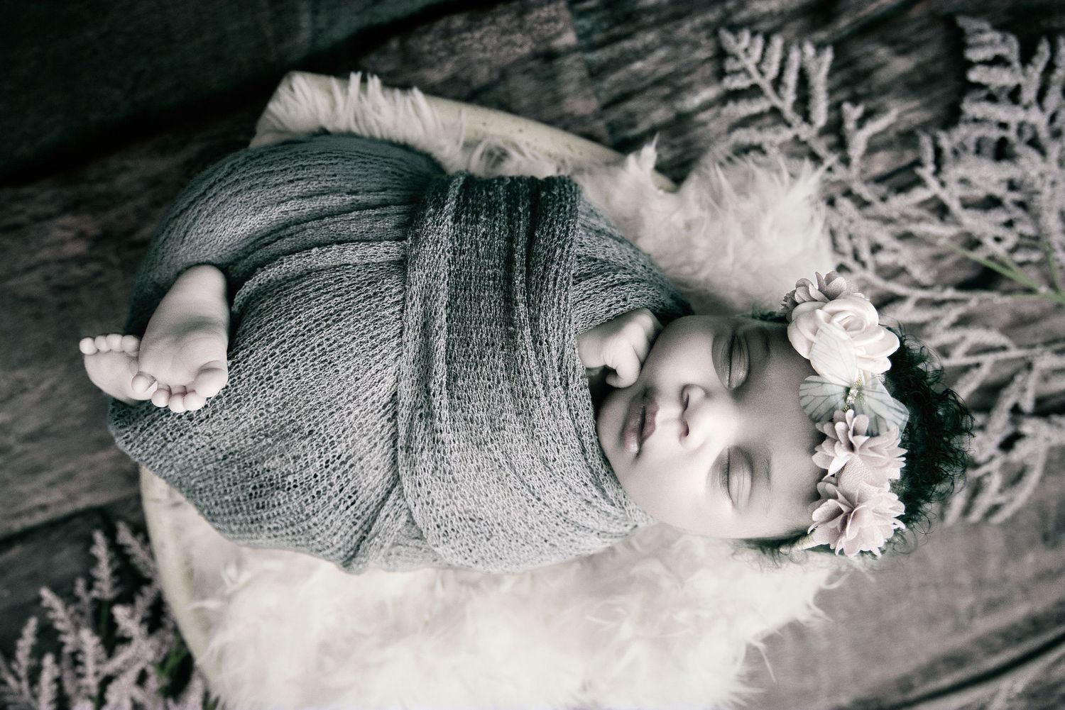 Black and white photo of a sleeping newborn baby wrapped in knit fabric with a floral crown on a fluffy white blanket.