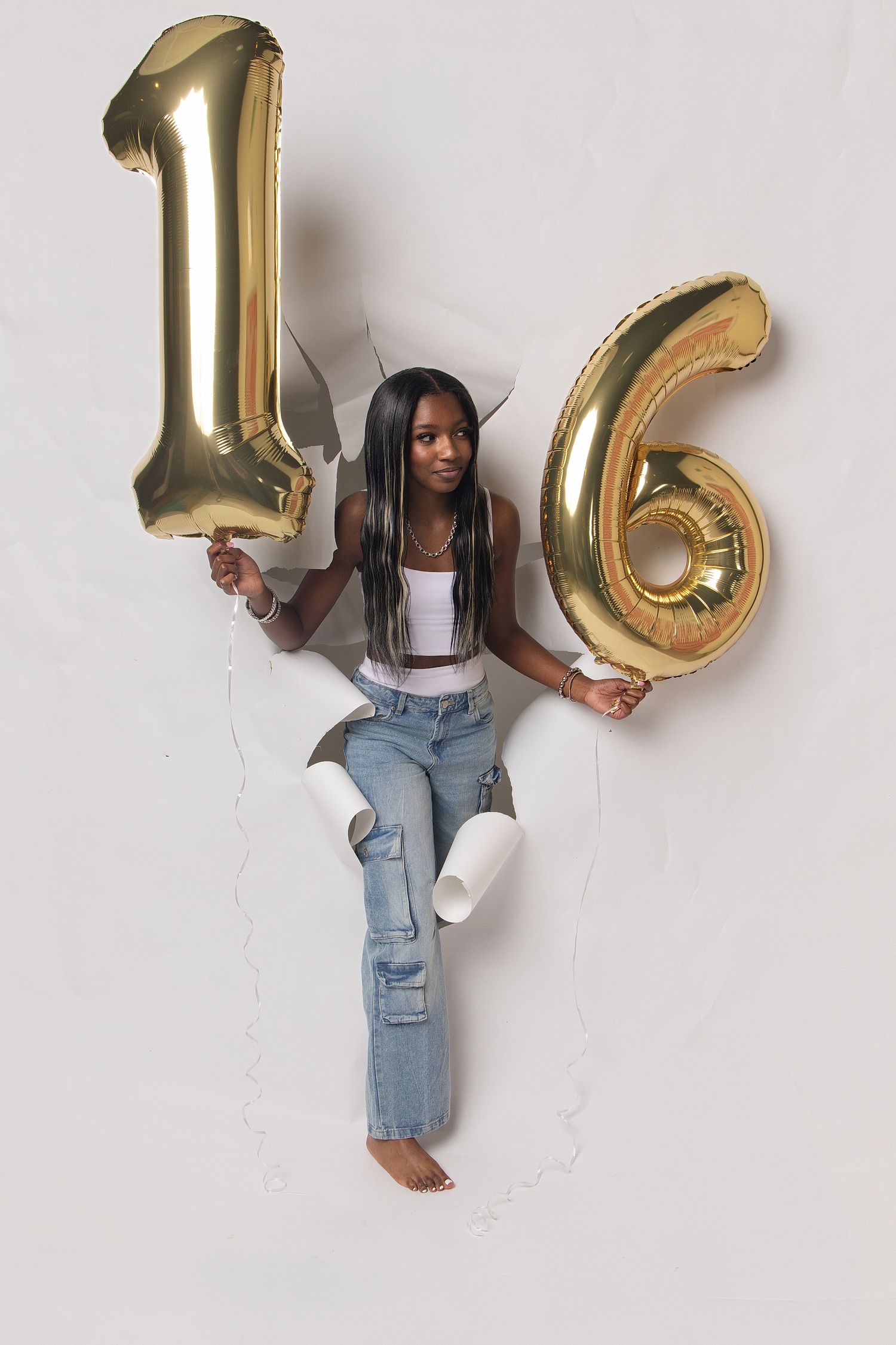 A young woman holds gold number 16 balloons against a white wall, wearing ripped jeans and a white crop top.