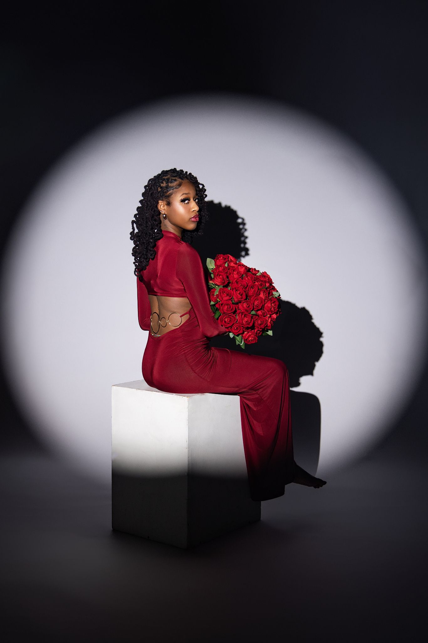 A figure in a red dress poses elegantly on a white cube against a dark background with dramatic lighting.