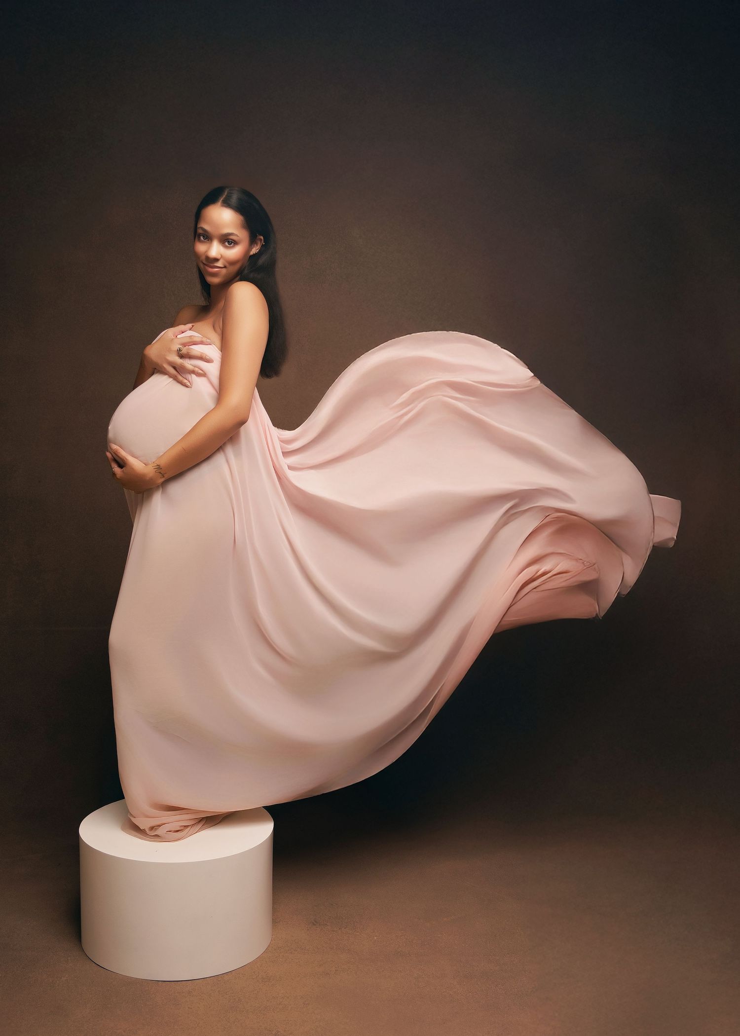 A graceful dancer in a flowing pink dress poses elegantly on a round white platform against a dark background.