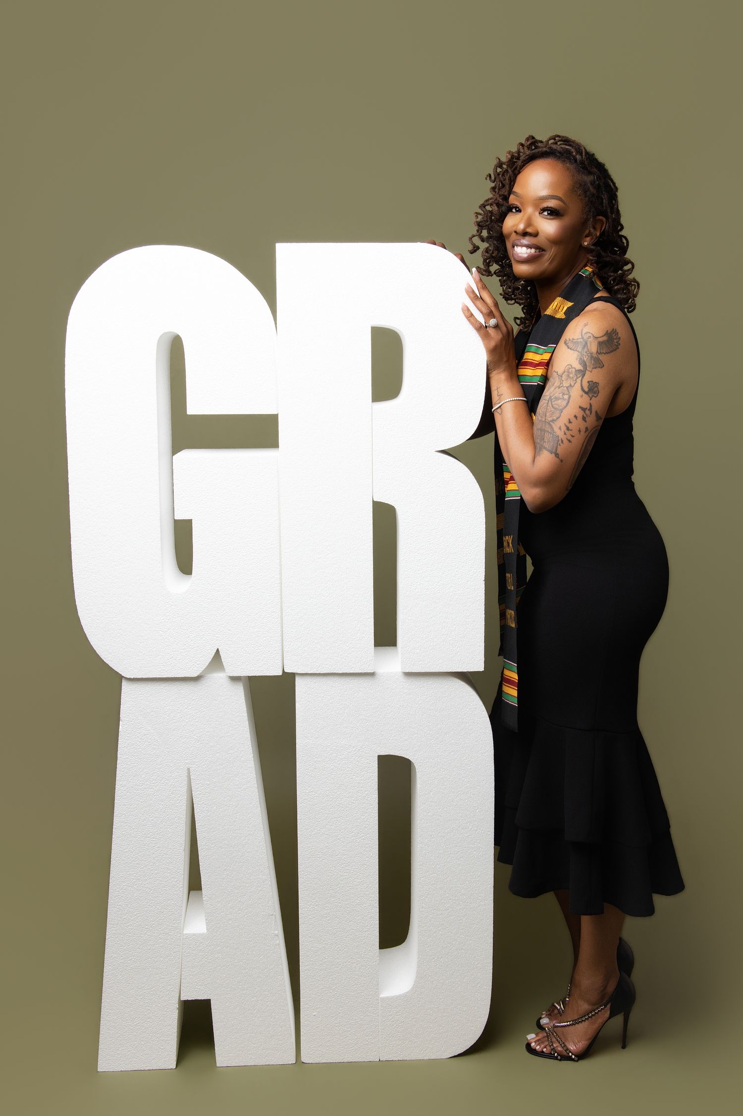 Woman in black dress posing proudly next to large white GRAD letters, celebrating her graduation achievement.