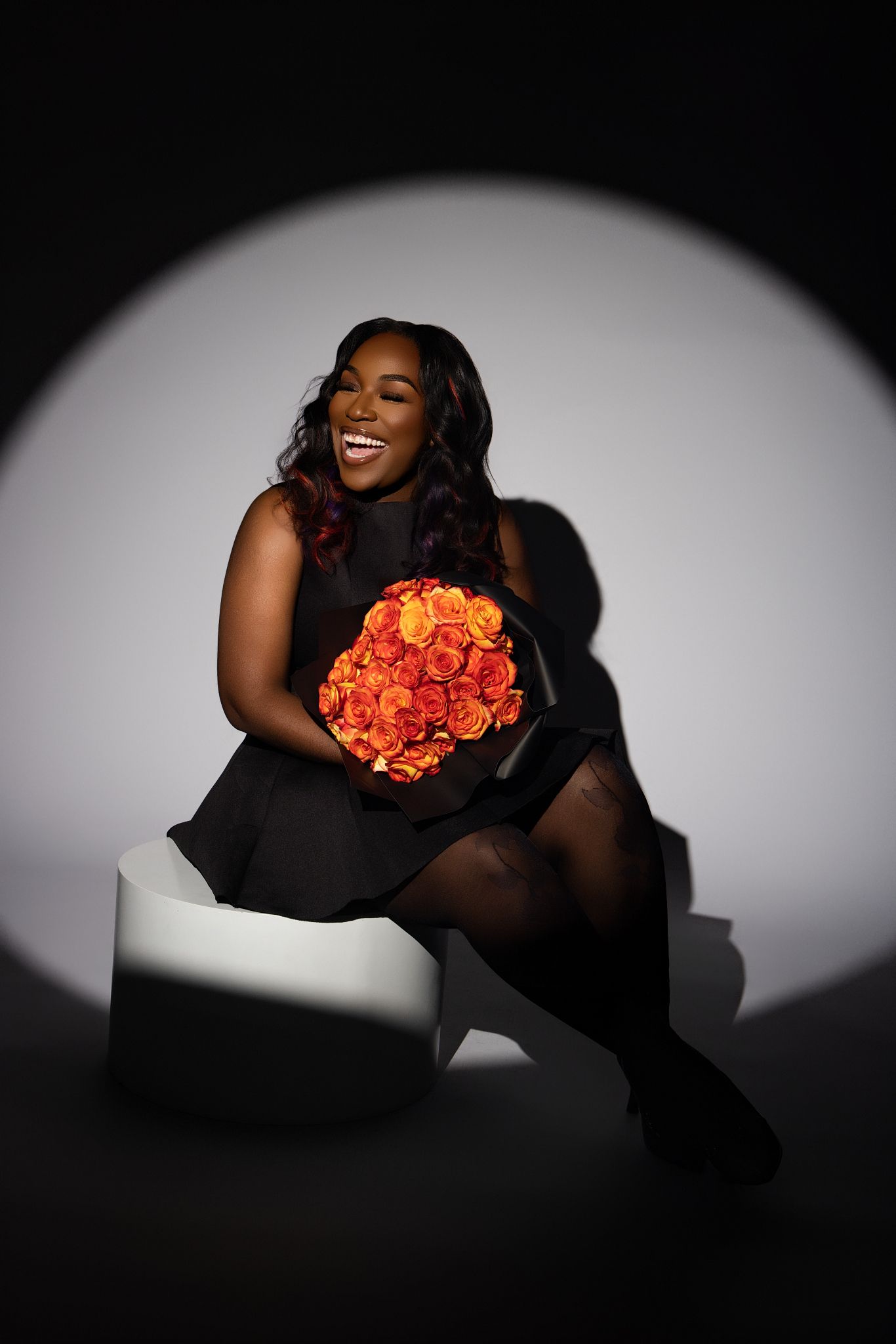 Elegant portrait against dark backdrop featuring person in black dress holding vibrant orange rose bouquet.