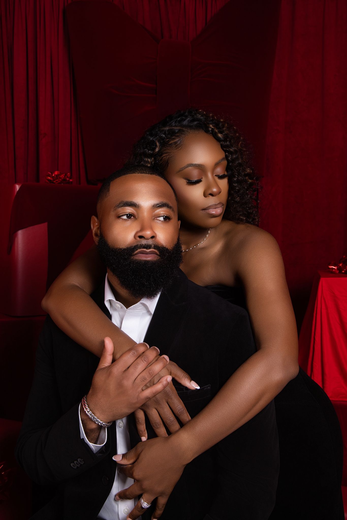 A romantic couple in formal attire embraces in front of deep red curtains during an intimate photo session.