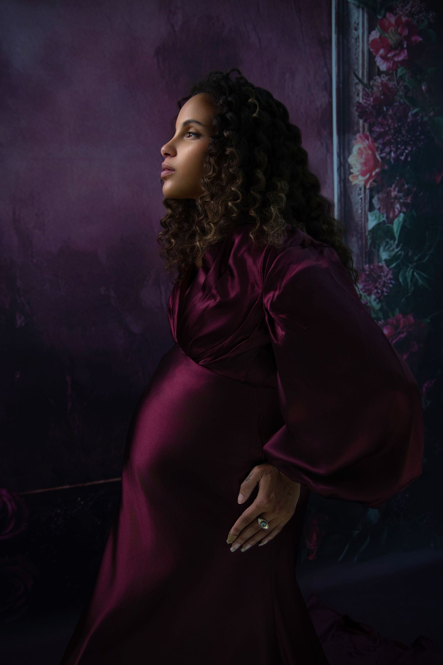 Woman in burgundy velvet dress poses dramatically against moody purple backdrop with floral accents.
