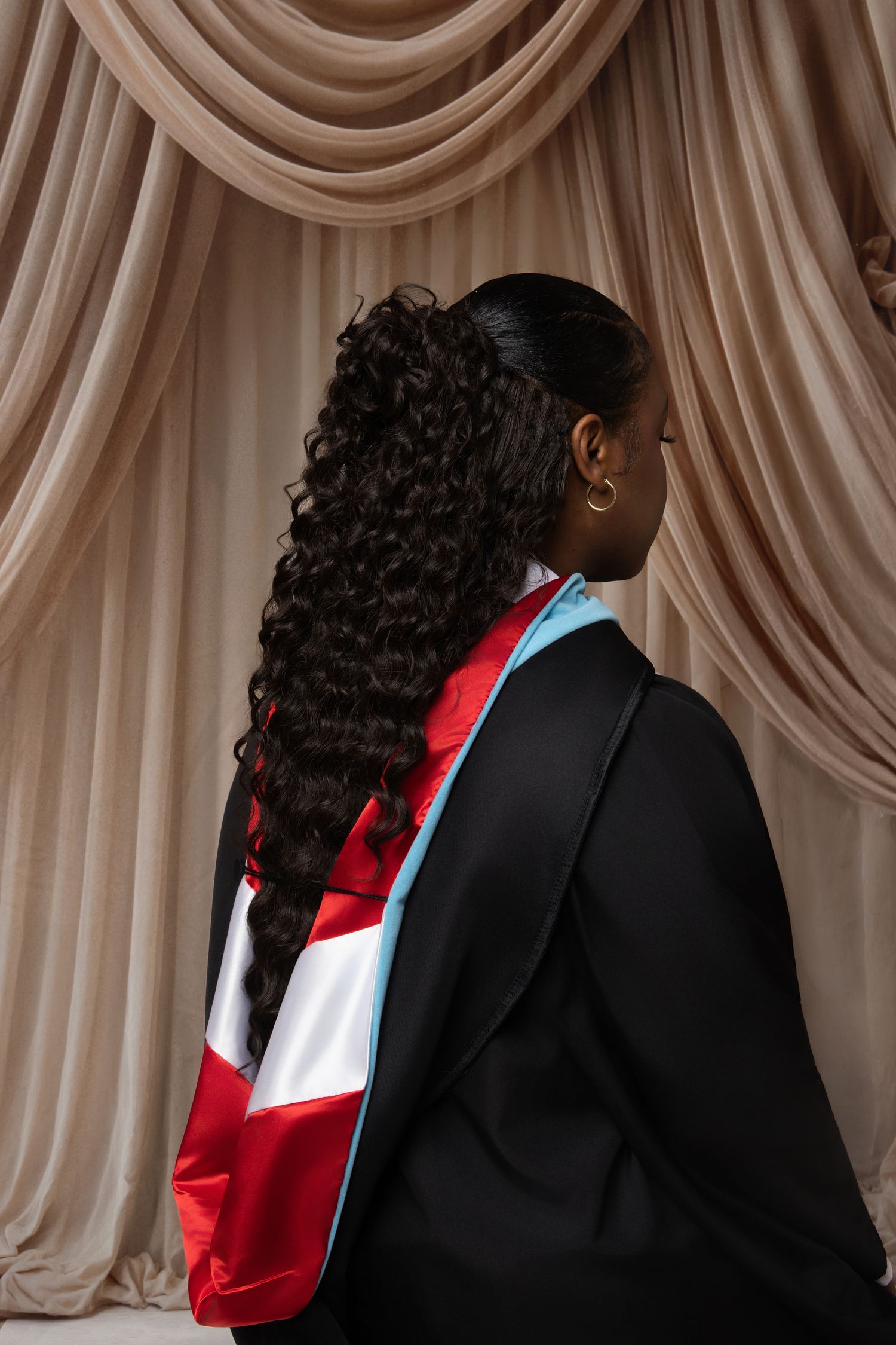 Black woman in graduation regalia with curly hair posing with back to camera in front of beige drapes.