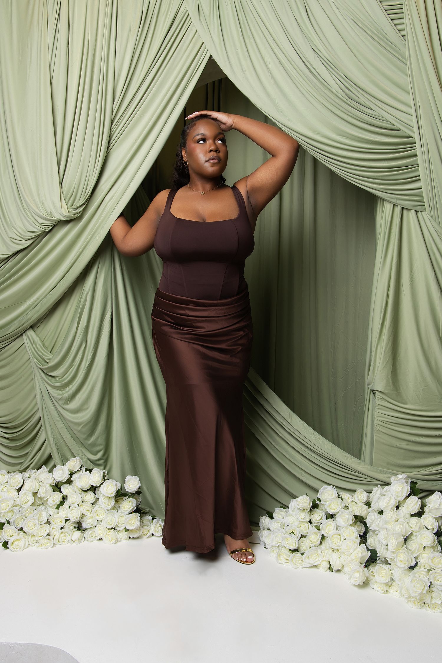 Woman in brown satin maxi dress poses elegantly among white roses with sage green draped curtains backdrop.
