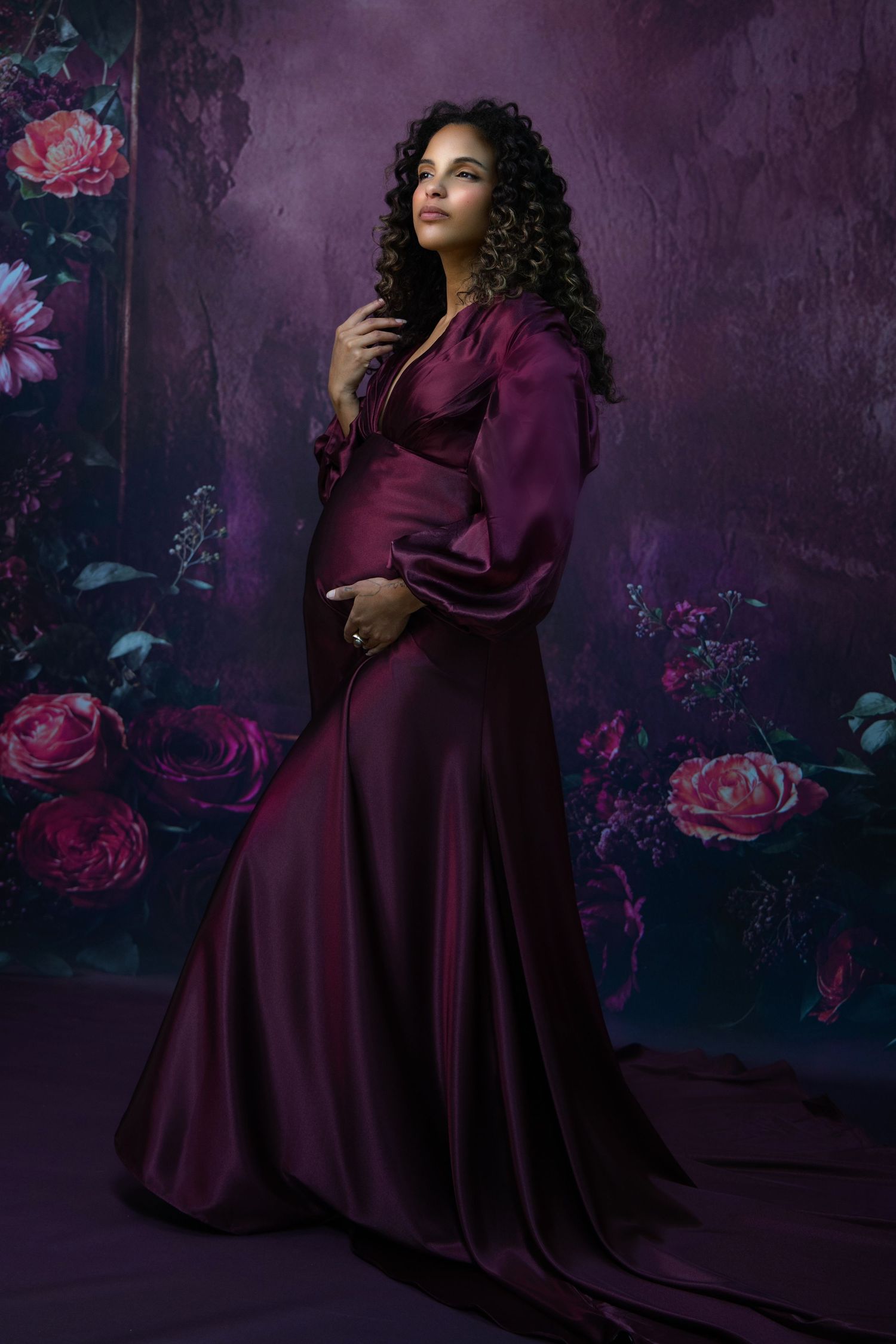 Elegant maternity portrait in flowing burgundy dress with dramatic lighting and floral background.