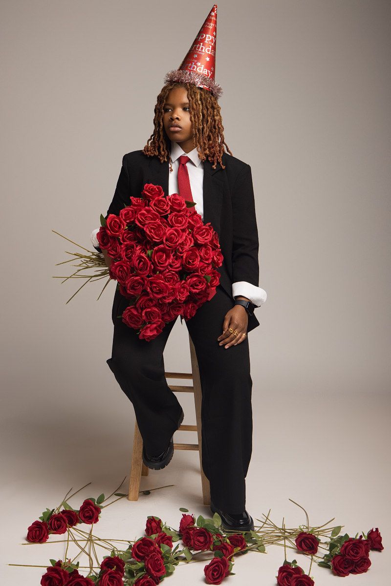 A figure in a black suit and party hat sits surrounded by scattered red roses on a light studio backdrop.