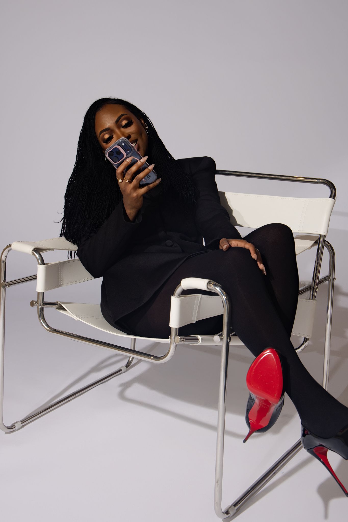 A figure in black attire poses dramatically on a sleek metal chair against a white background.