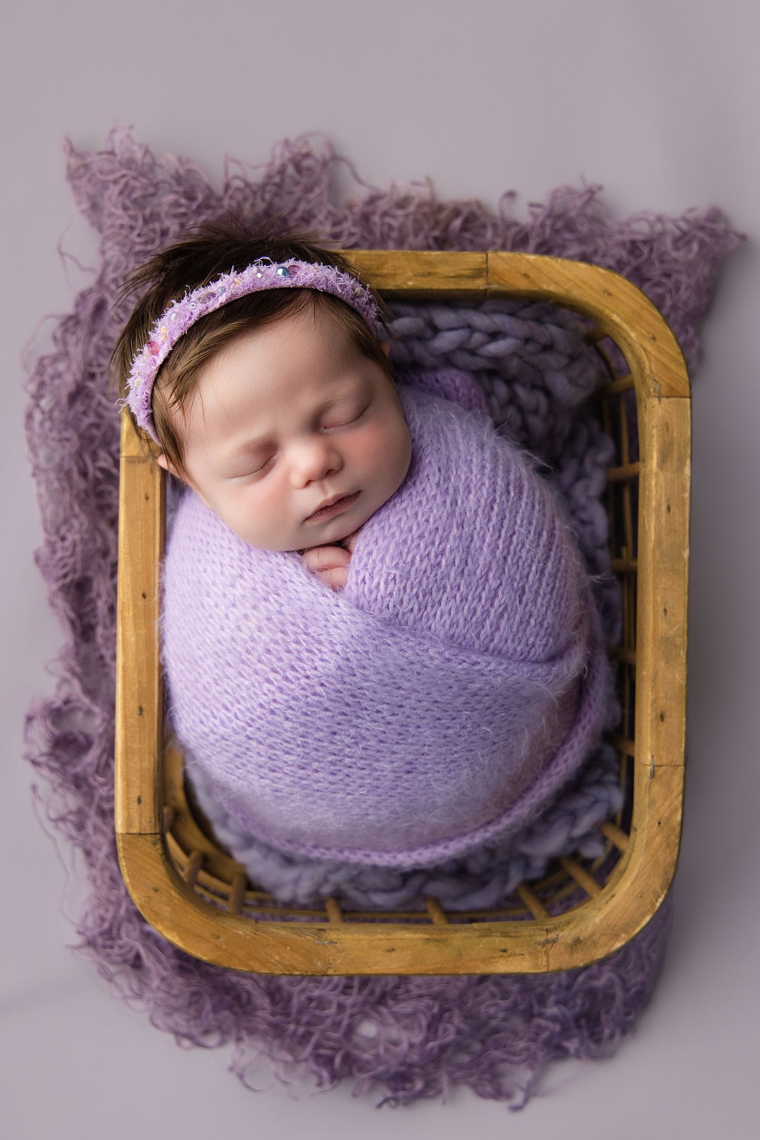 Newborn baby girl swaddled in lavender knit wrap with floral headband, sleeping peacefully in a wooden basket.