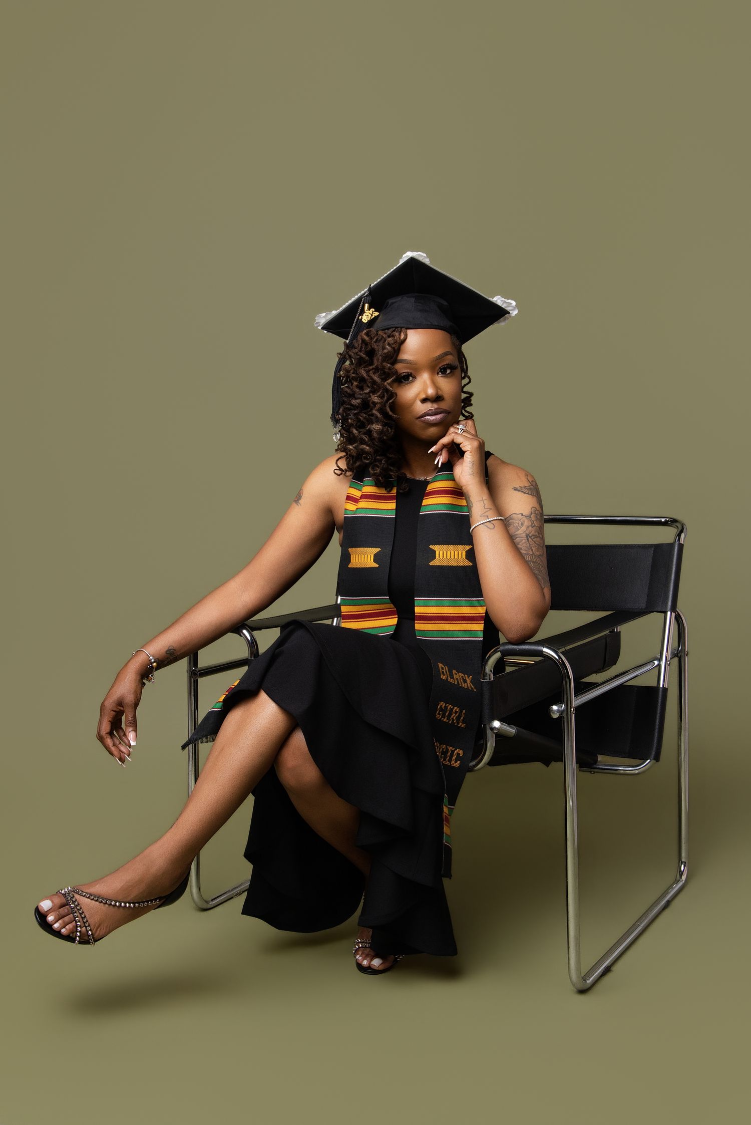 Young woman in graduation cap and kente stole poses in black dress on modern chair against olive background.