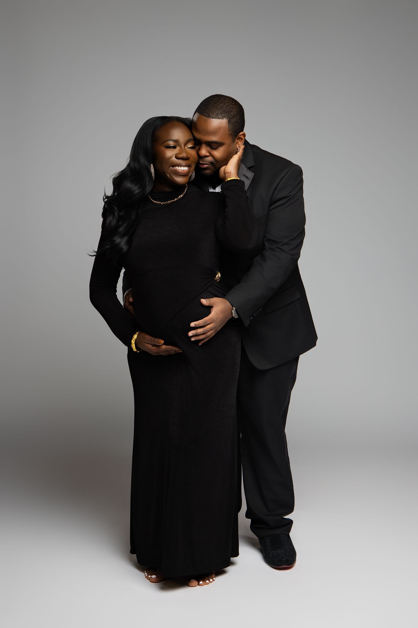 An intimate maternity portrait of a couple in black formal attire against a gray studio backdrop.