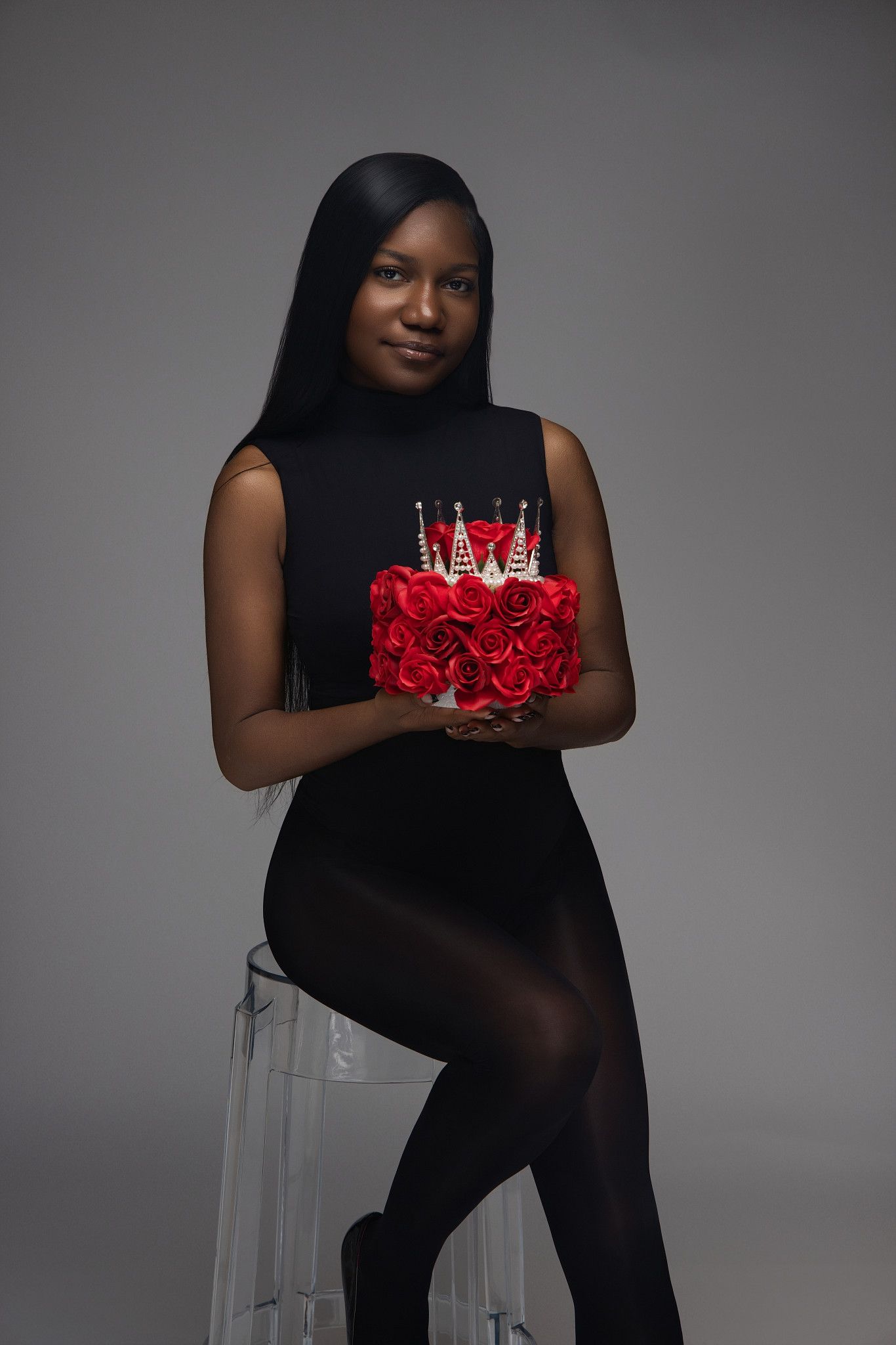 Elegant woman in black dress poses with red rose birthday cake on clear stool against gray backdrop.
