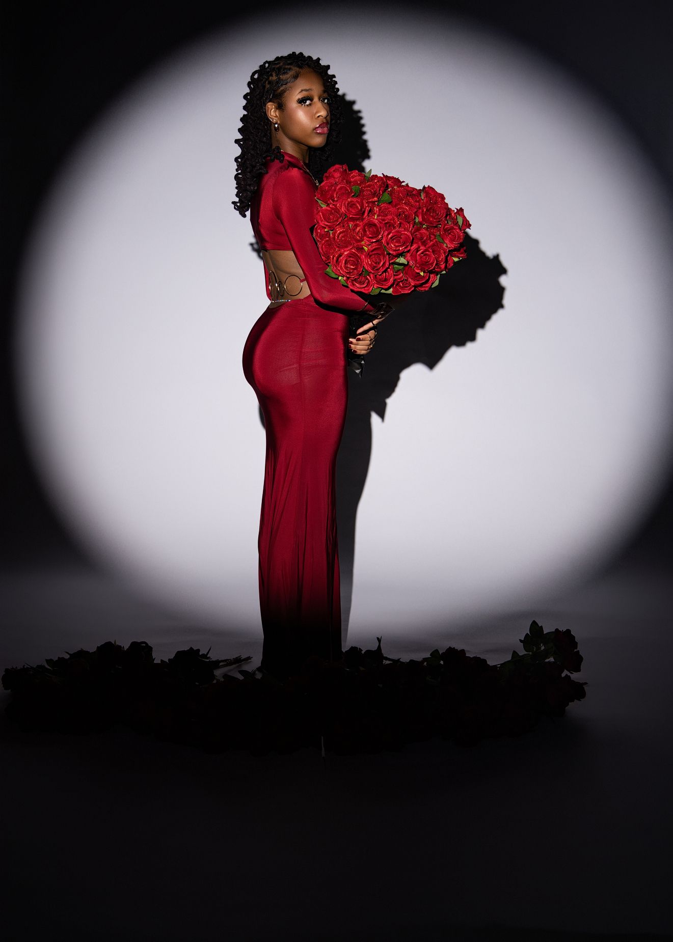 Figure in a stunning red gown holds a luxurious bouquet of red roses against a dramatic spotlight backdrop.