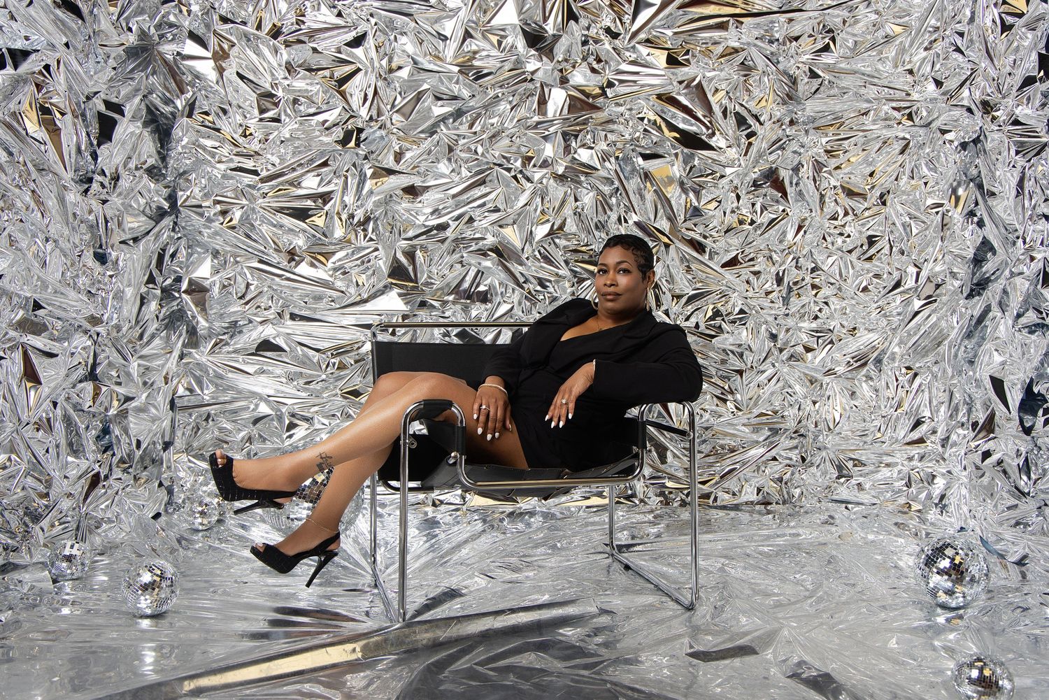 Woman in black dress poses confidently on chrome chair surrounded by silver metallic sculptural wall installation.