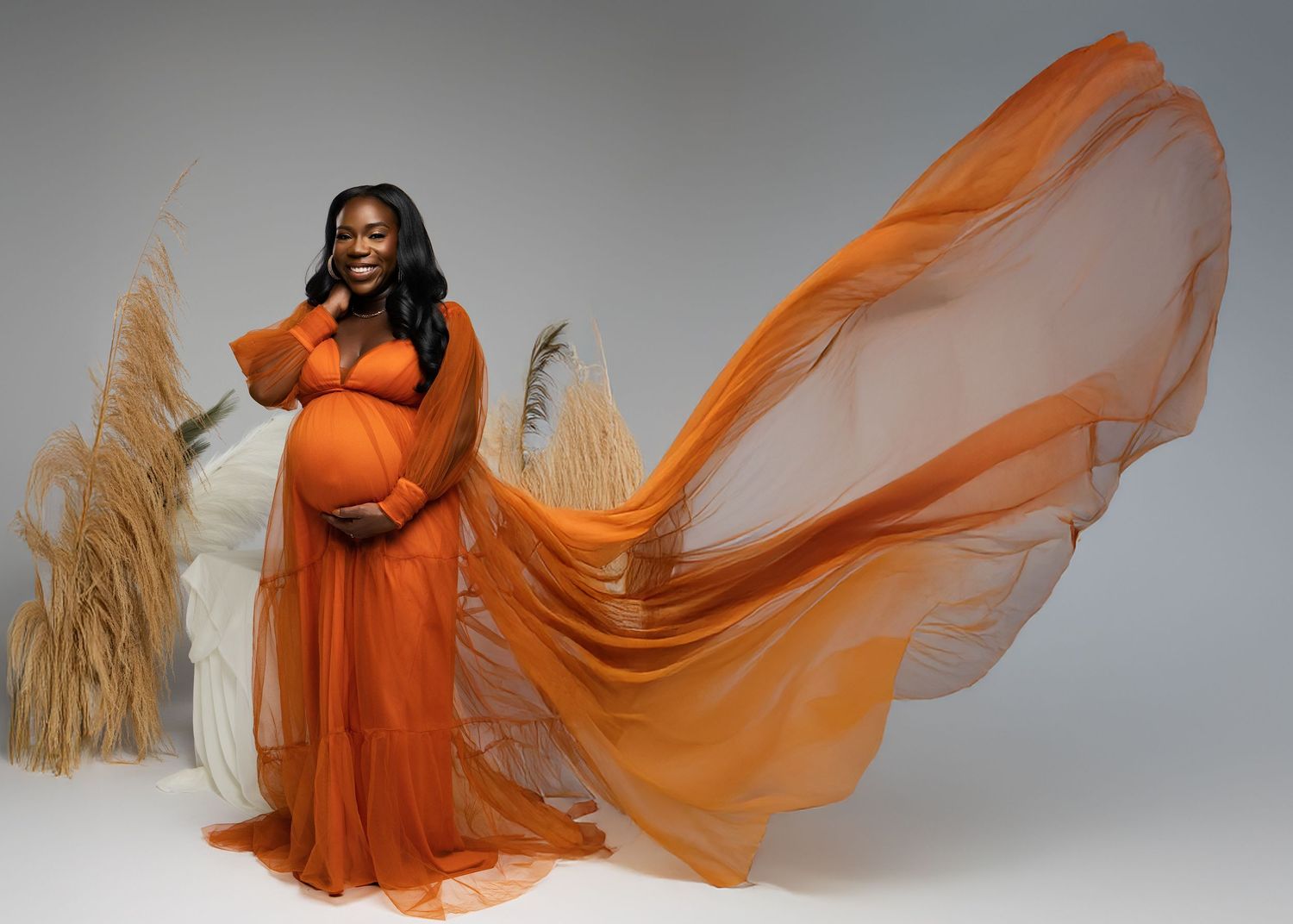 Elegant maternity photo featuring a flowing orange dress with billowing fabric against a light backdrop with pampas grass.