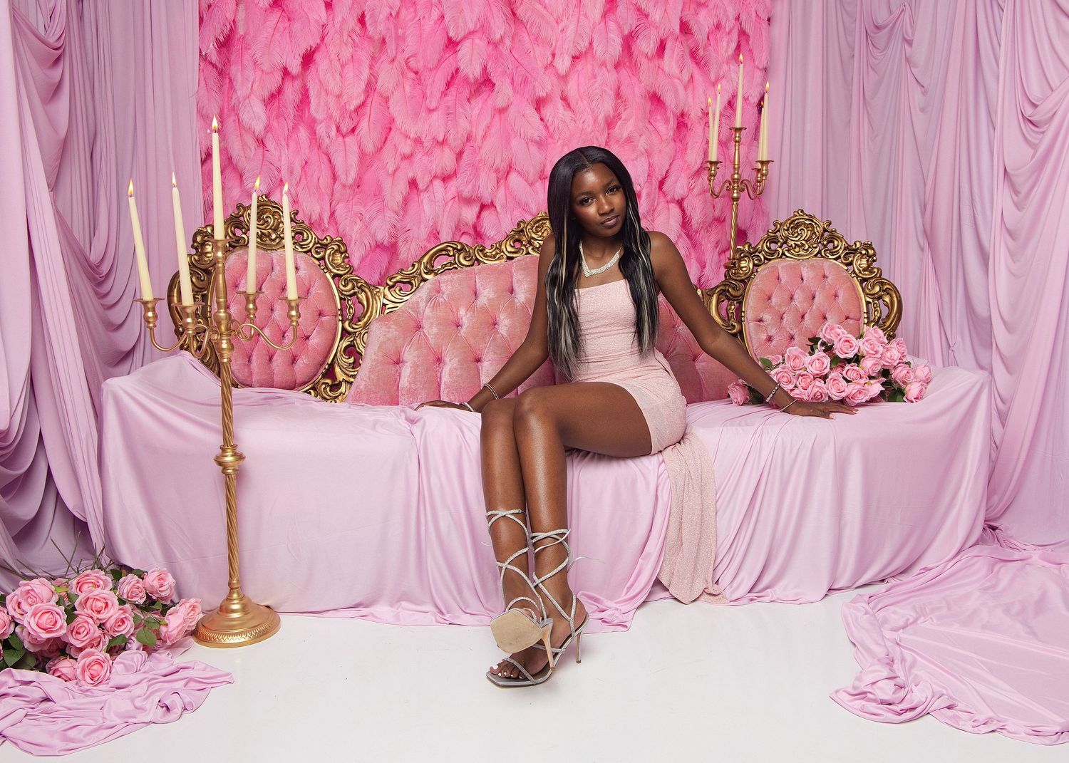 Young woman in pink dress poses on ornate gold sofa with candelabras and pink floral backdrop.