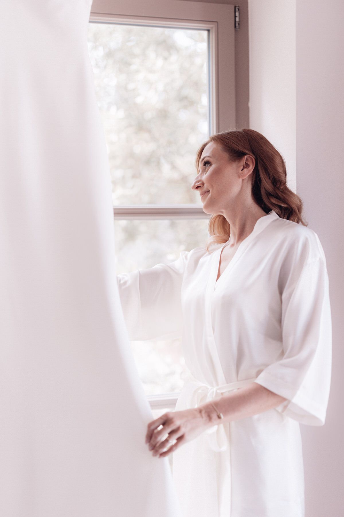 Bride standing by the window in a robe, quiet and intimate wedding morning moment