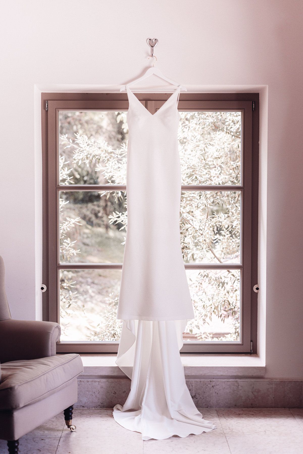 Minimalist wedding dress hanging in front of a window, natural light and elegant bridal details