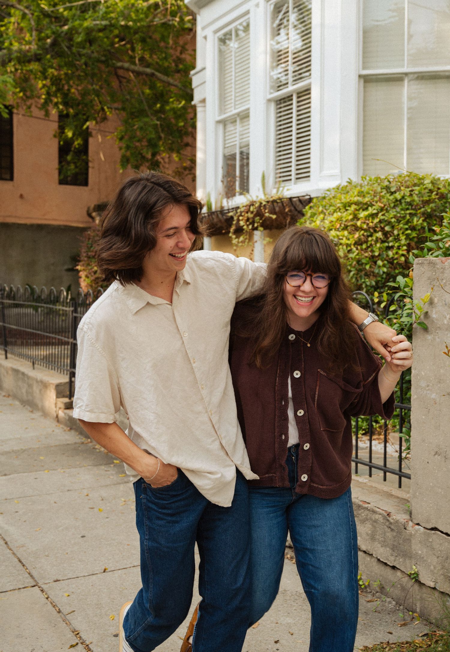 Two friends laughing and walking together on a sunny urban sidewalk, wearing casual shirts and jeans.