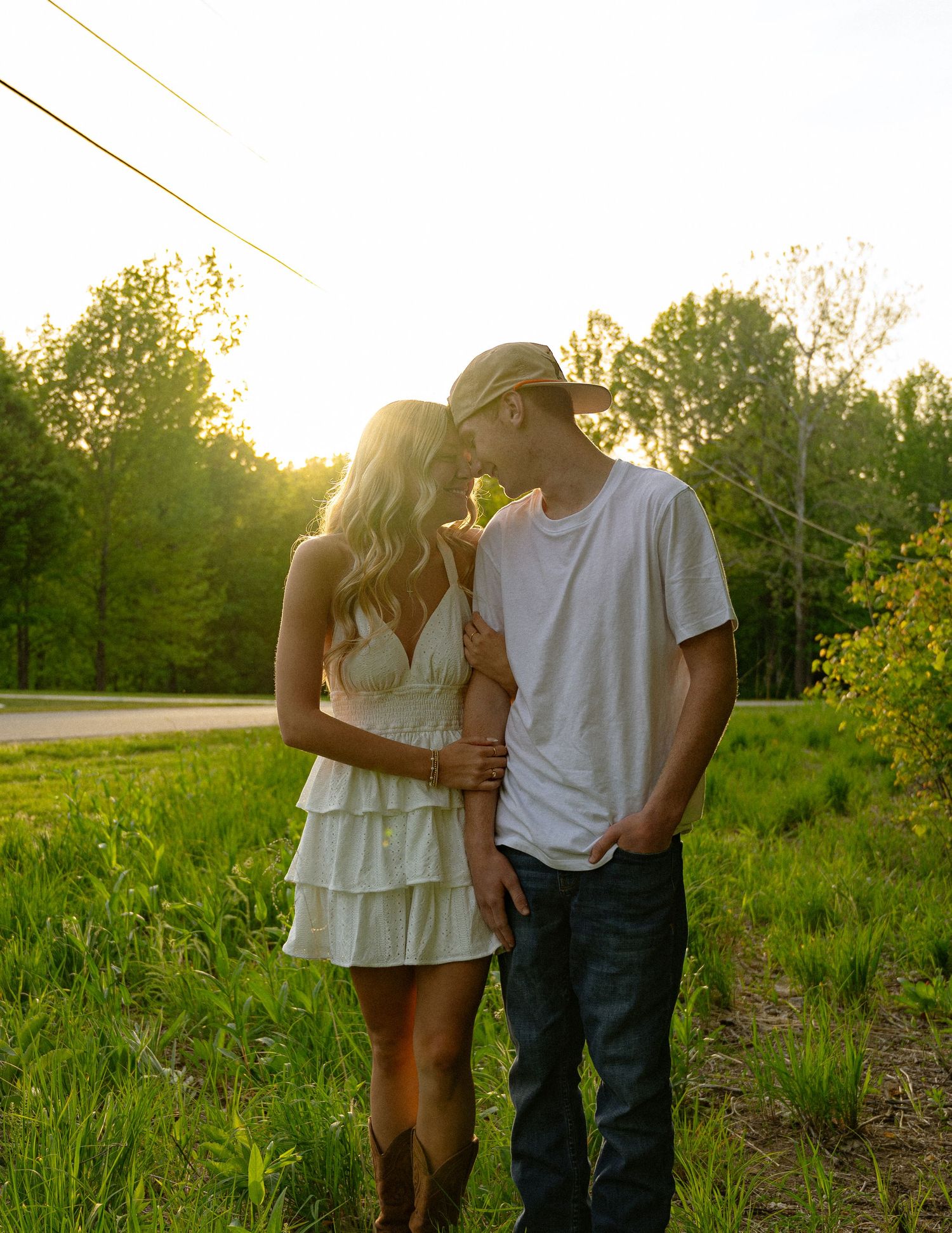 A couple shares a romantic moment at golden hour in a lush green field, woman in white dress and man in hat.