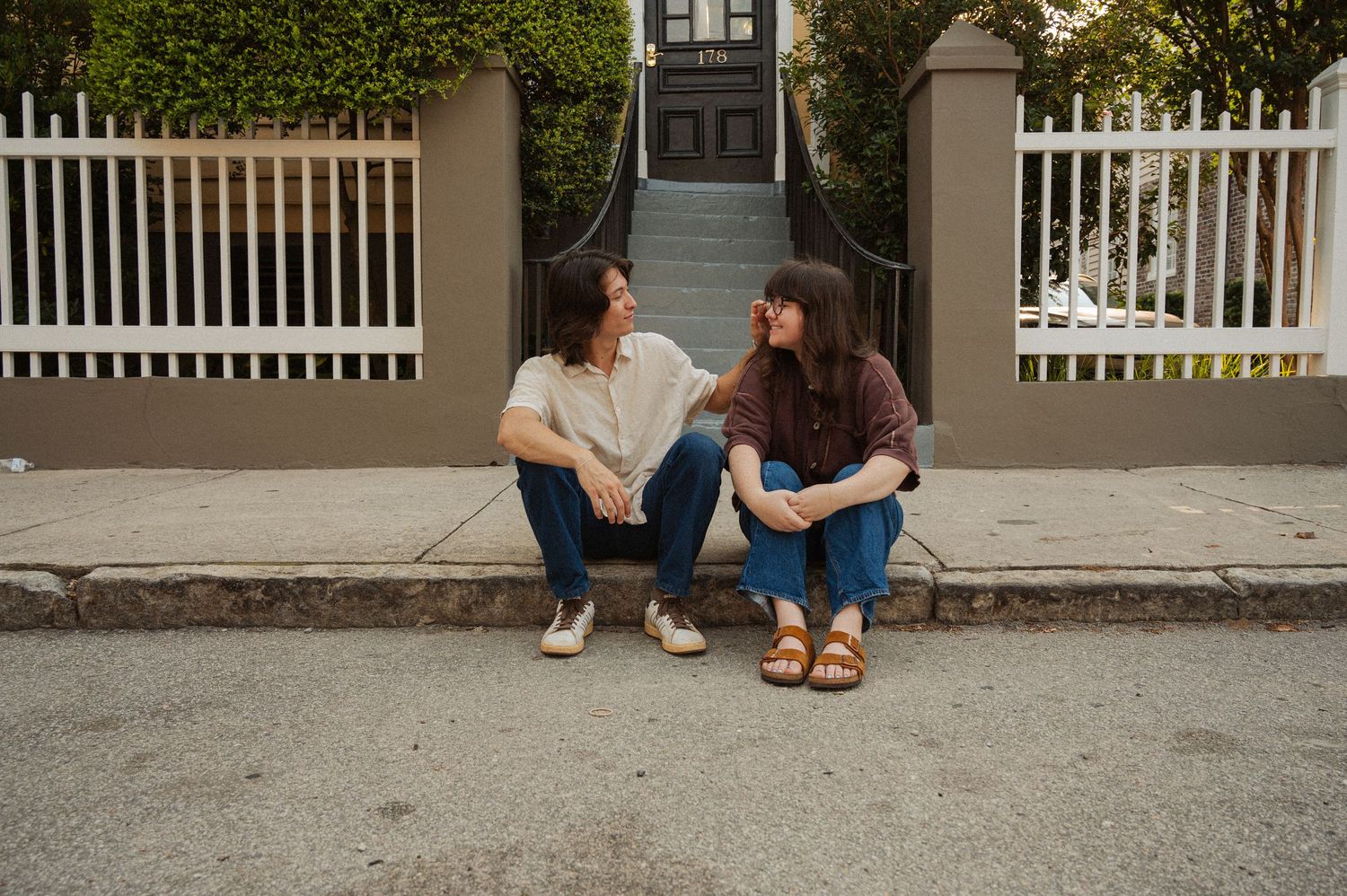 Two young women sit on a curb chatting in front of a white fence and steps outdoors.