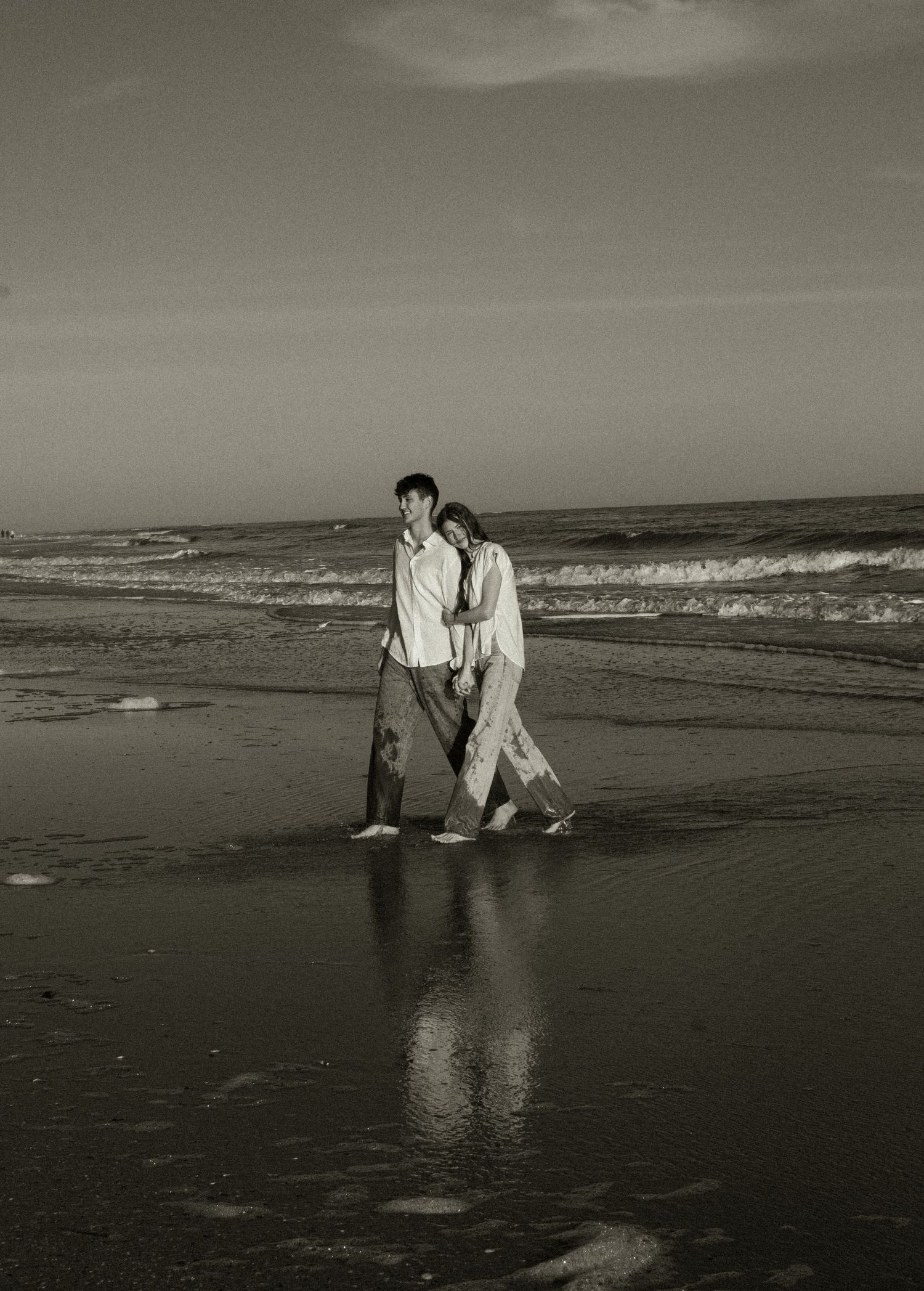 A couple in white clothing stands together on a dark sandy beach at sunset creating reflections in the wet sand.