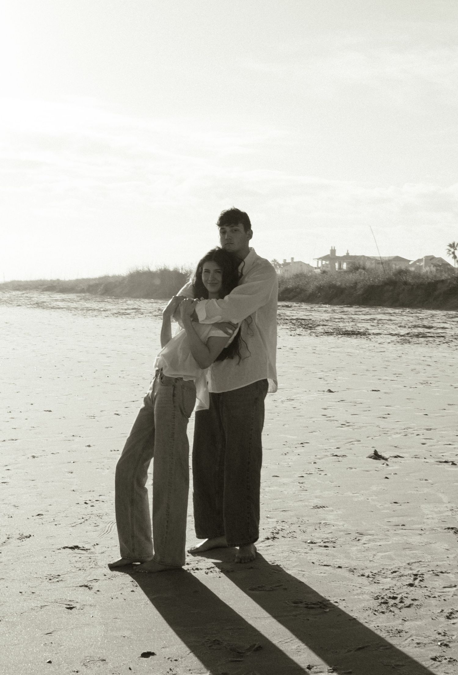 Black and white photo of a couple embracing on a sandy beach with ocean waves and coastal homes in the background.