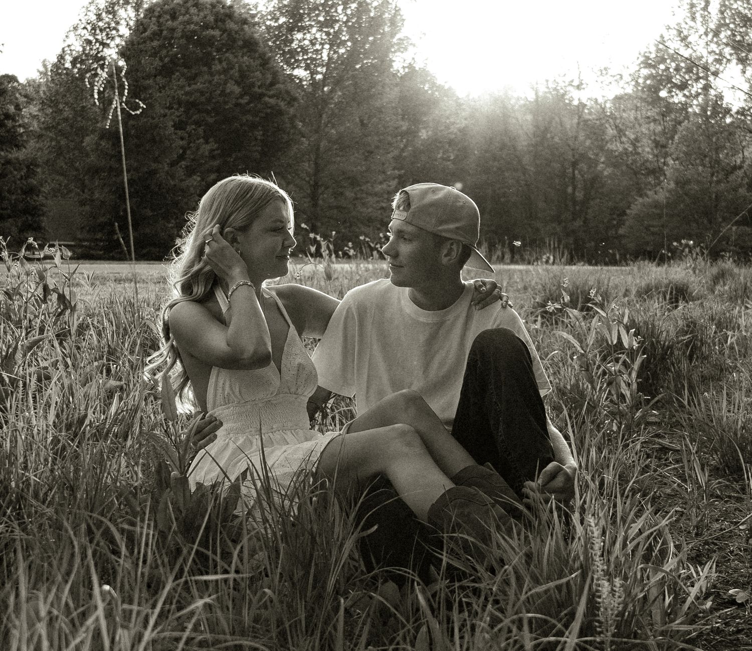Black and white photo of a young couple sitting together in a grassy field with sunlit trees in the background.