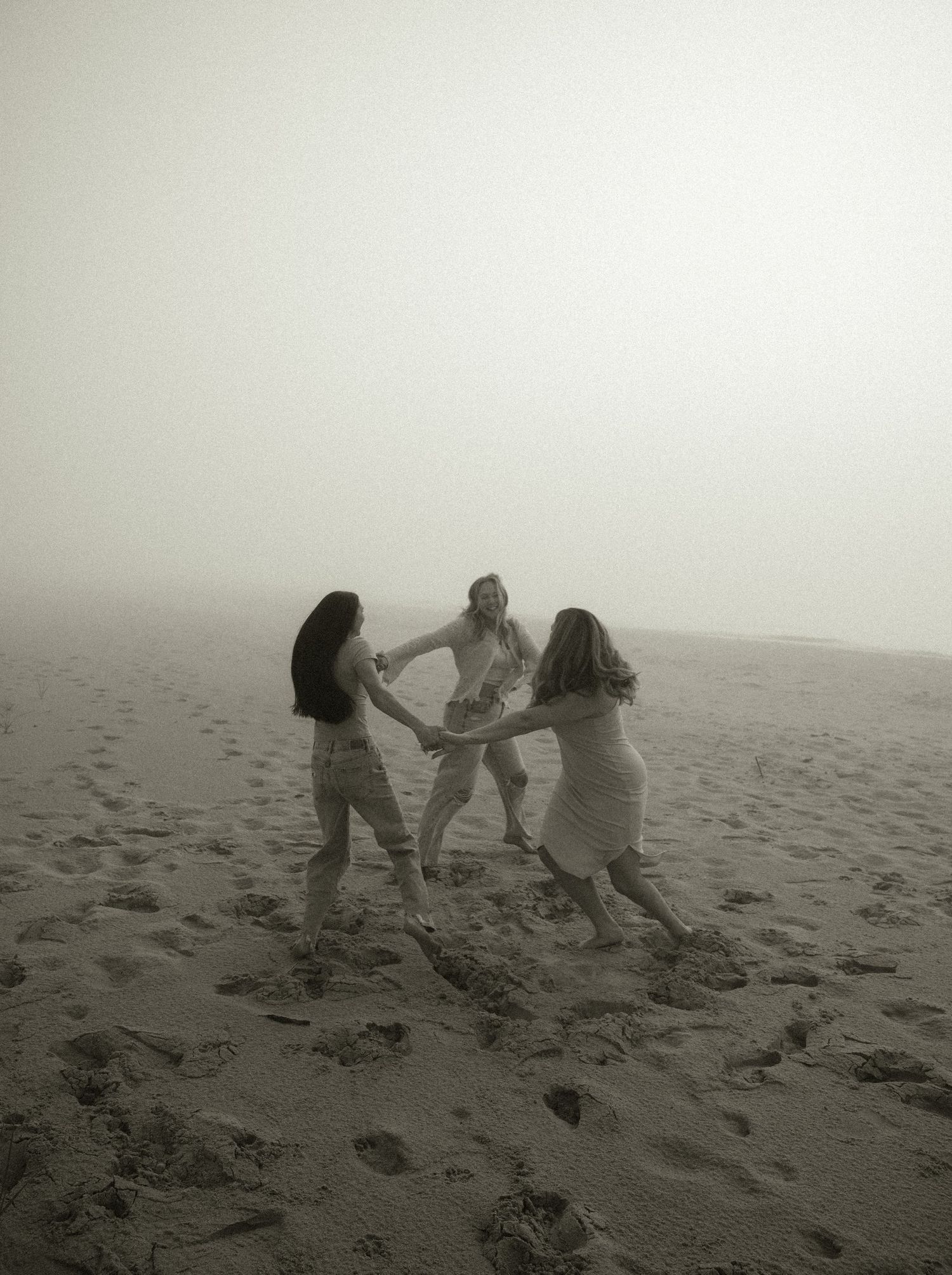 Three women holding hands in a circle on a foggy beach, dancing together on sandy shore in black and white photo.